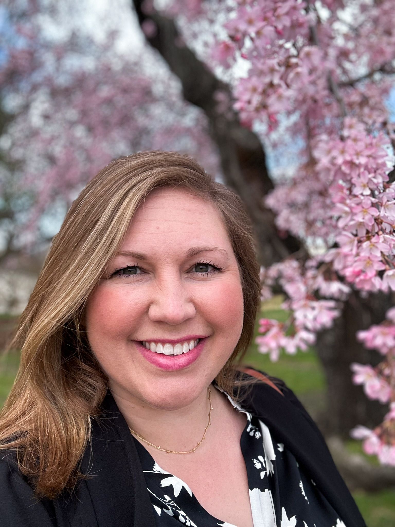 Woman smiling in front of a blooming pink tree, wearing a black blazer and patterned top.