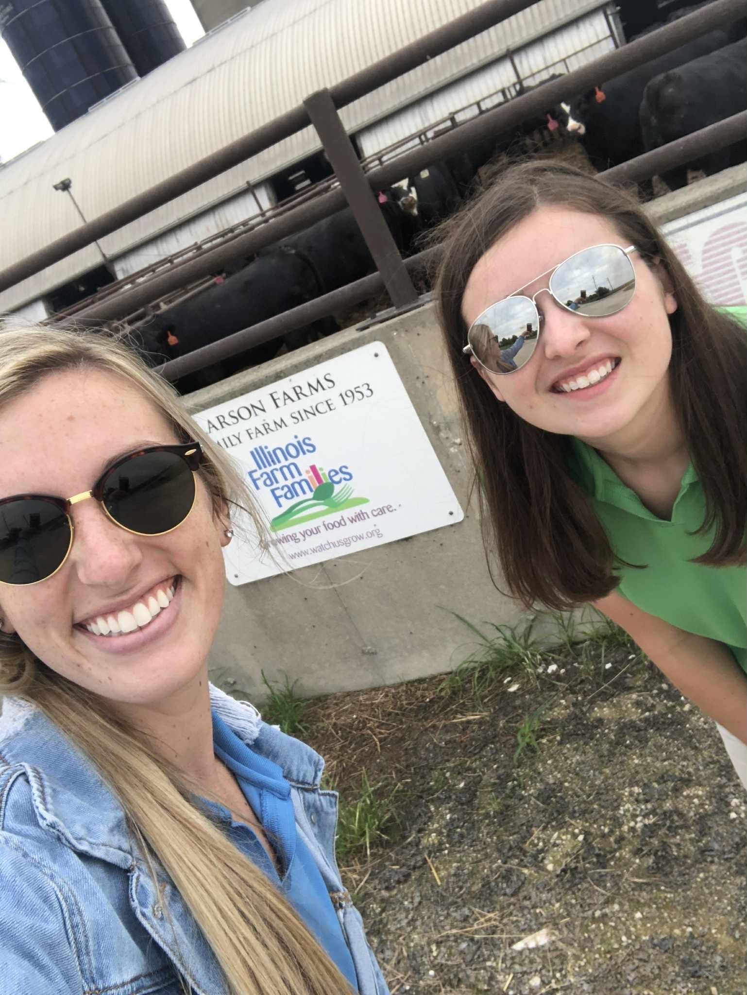 Two smiling women in sunglasses, near a sign and building. One wears green, the other denim.