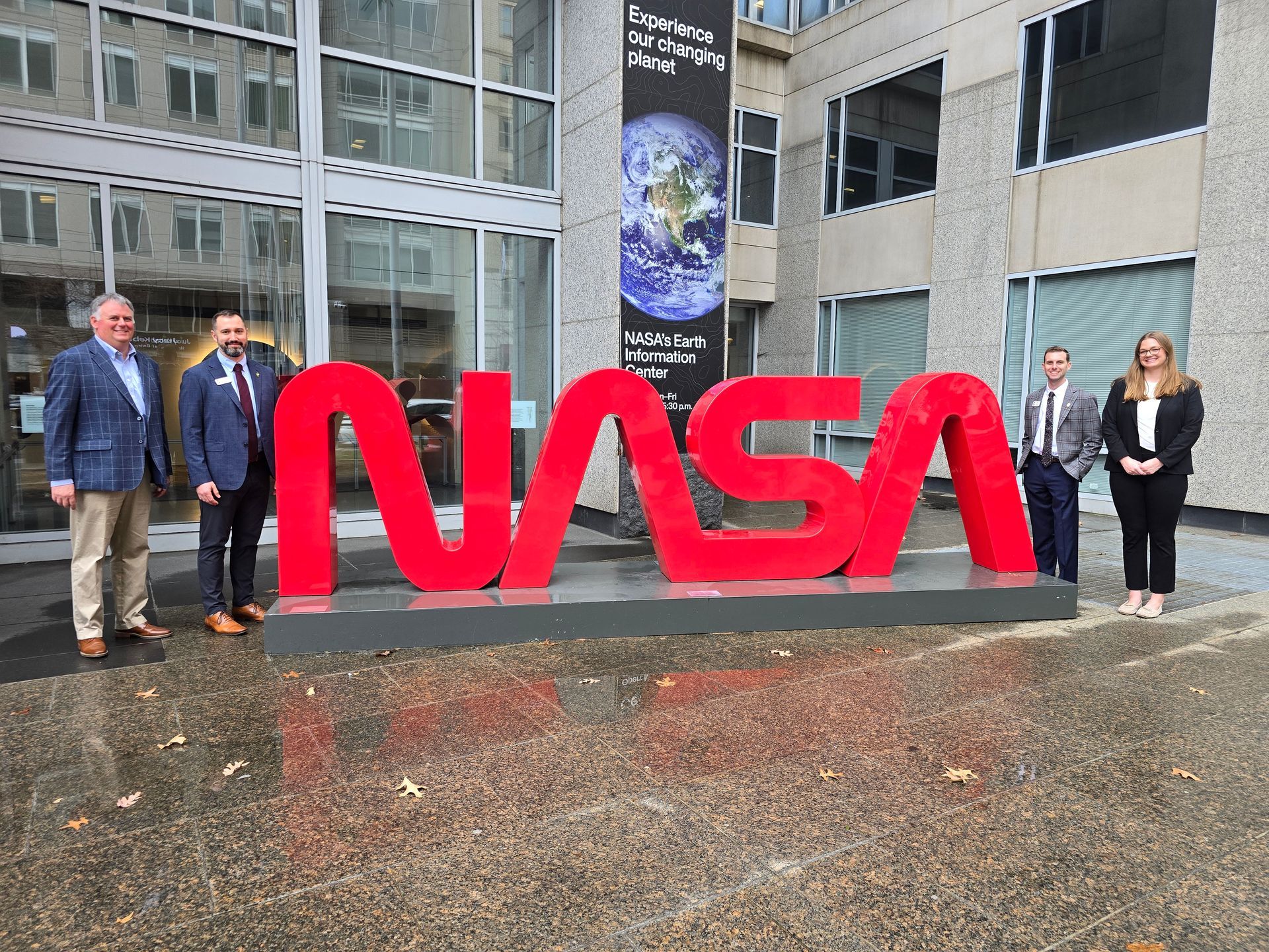 Four professionals stand near a large, bright red NASA logo sculpture outside a building with an Earth-themed banner.