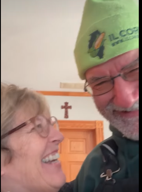 Smiling couple, woman with glasses, man in green hat, indoors, cross on wall behind them.