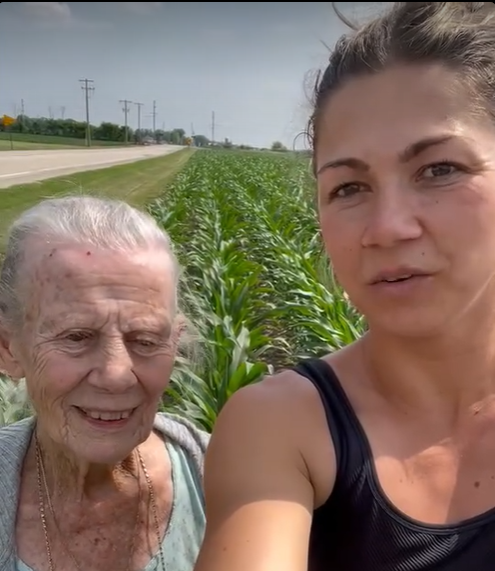 Woman and elderly person standing near a cornfield and road; person on right talking.