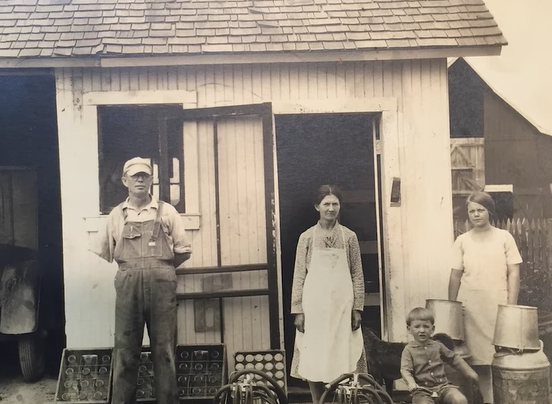 Family posing in front of a small building, eggs in front. Man in overalls, woman in apron, child seated.