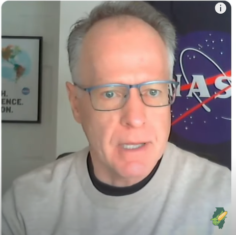 Man with glasses speaking, NASA logo in background, Earth map poster.
