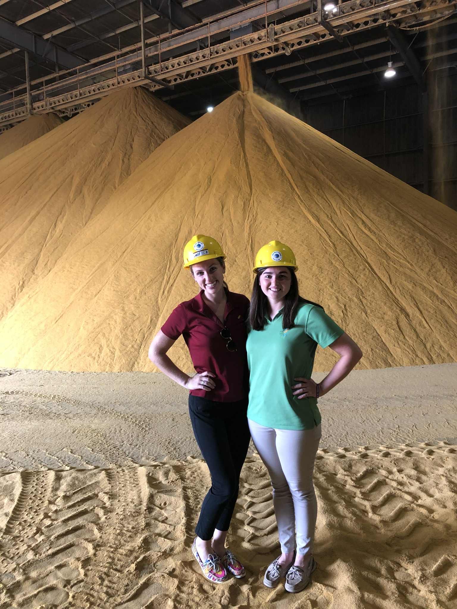 Two women in hard hats stand near large piles of grain.