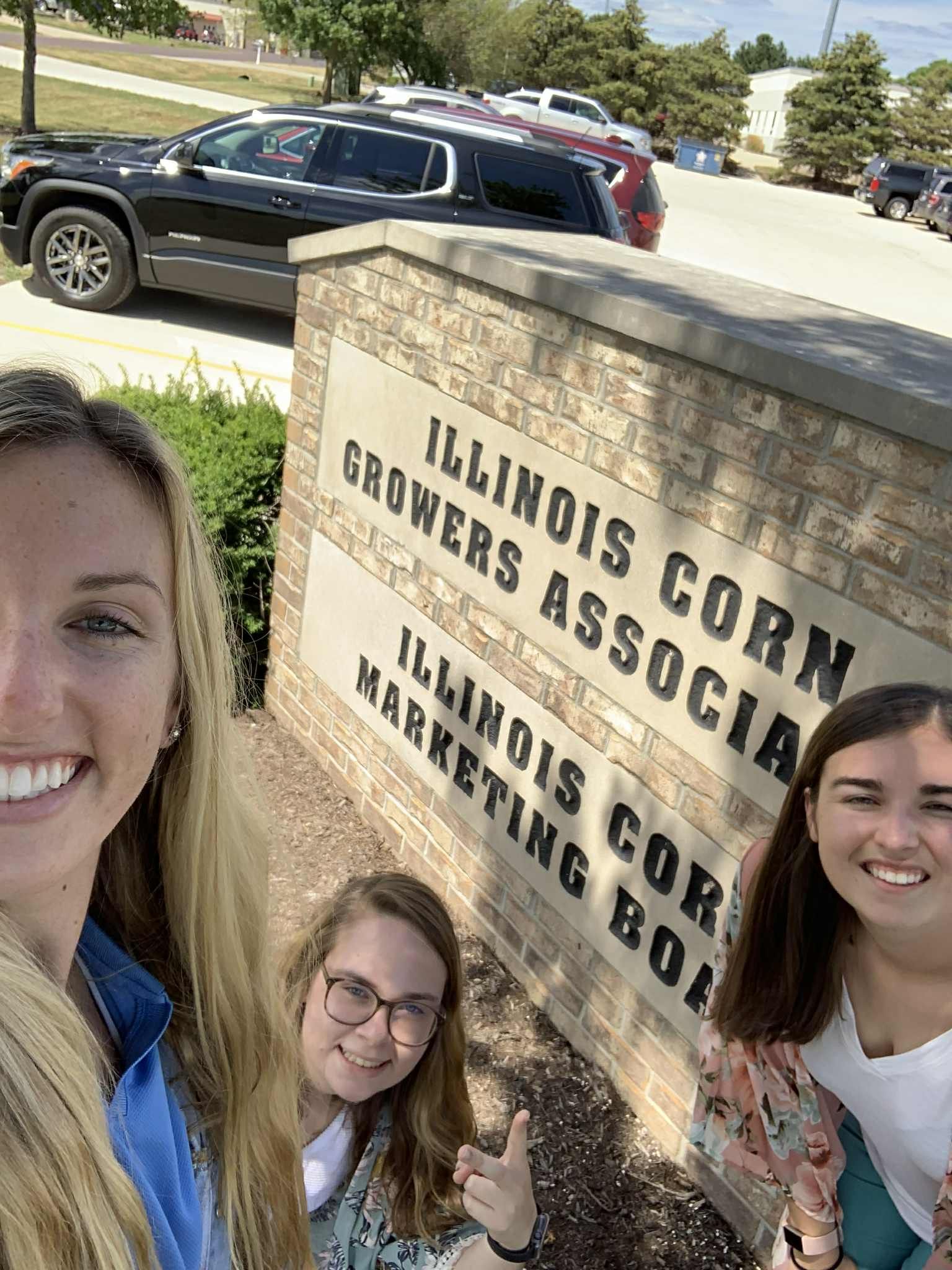 Three women pose in front of an Illinois Corn Growers sign. Sunny day, smiles and thumbs up.