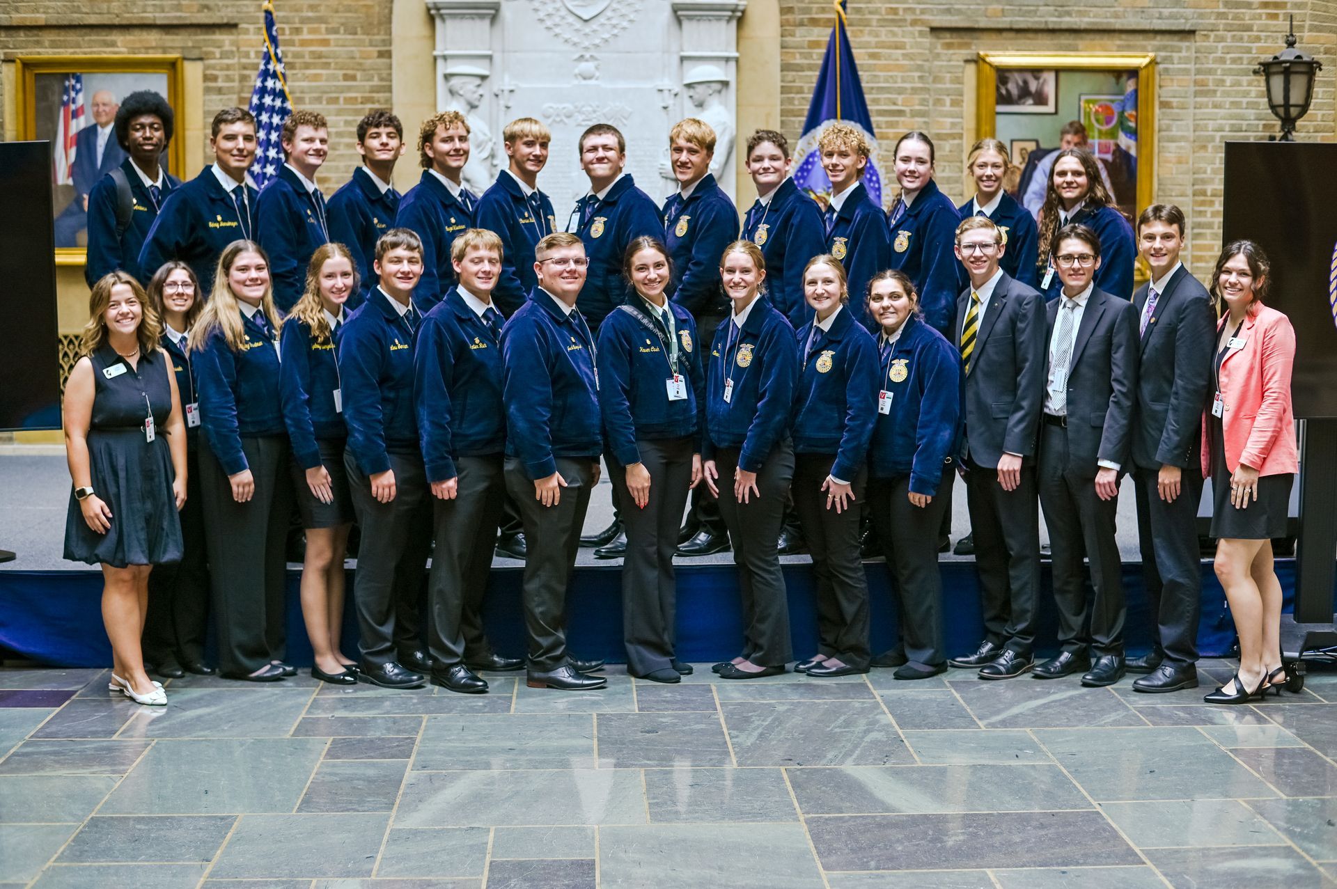 Group of FFA members posing for photo, some wearing jackets. Formal setting with flags and portraits.