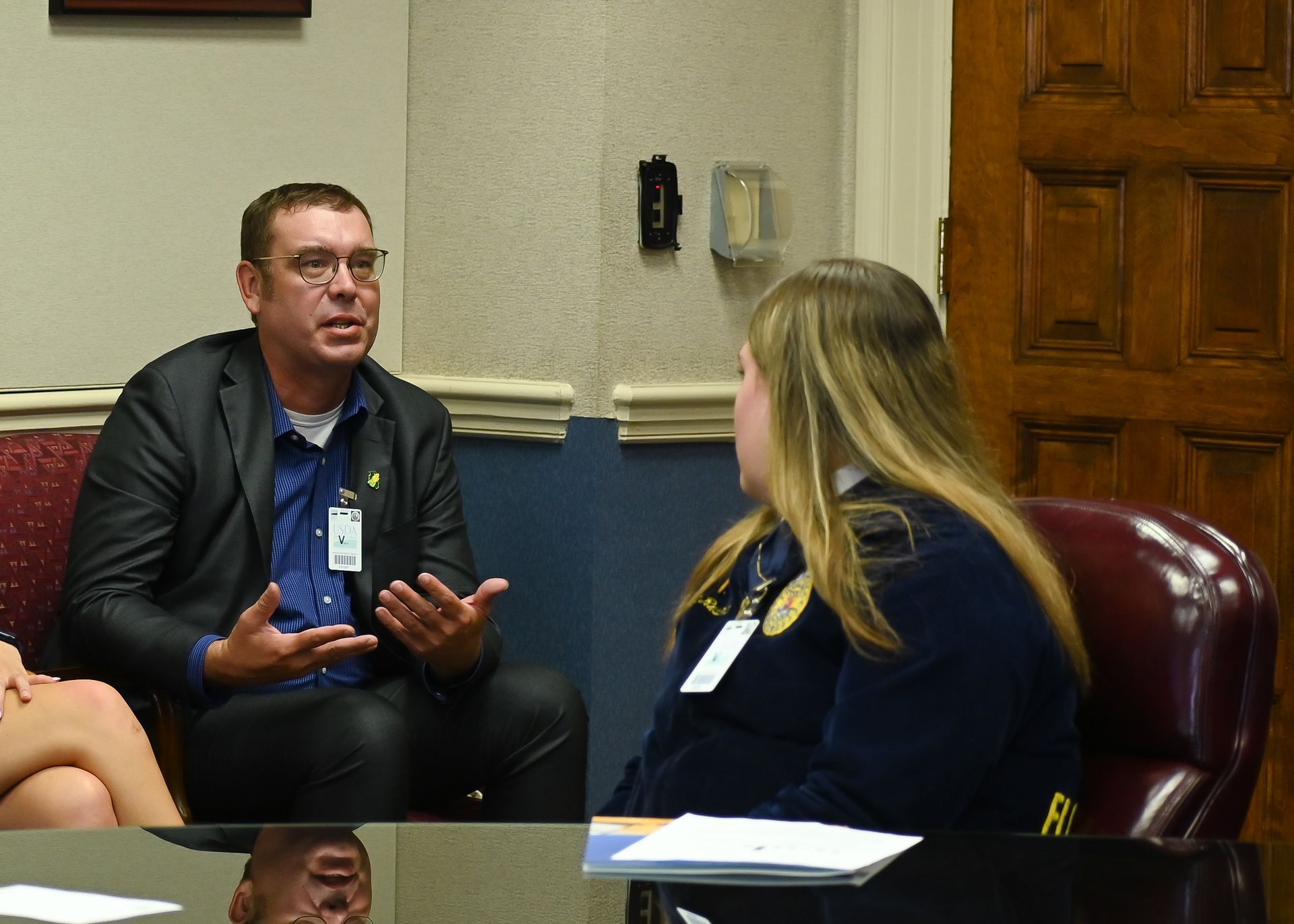 Man in blazer speaks with a woman in uniform, both seated. Interior shot, discussing.