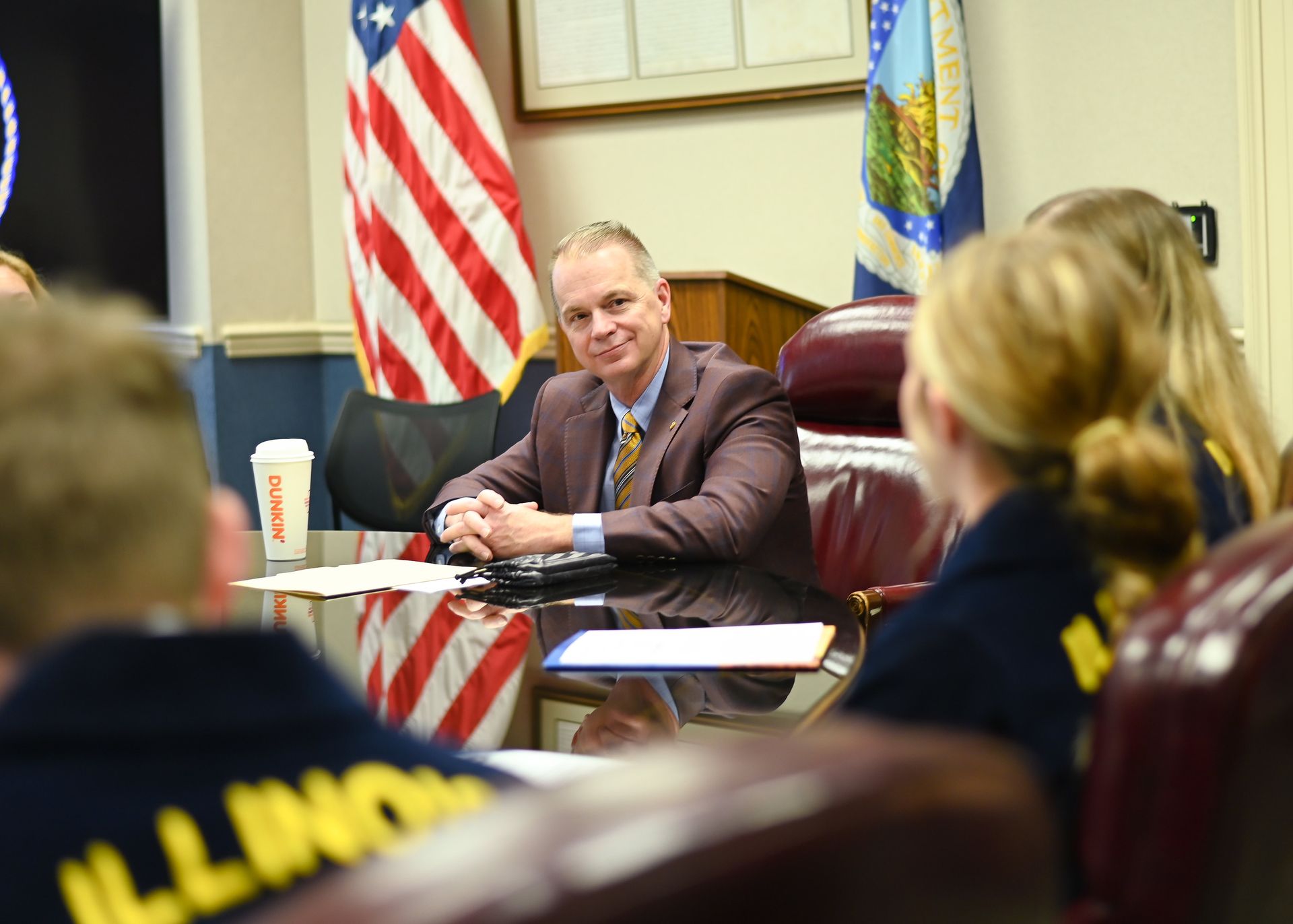Man in suit smiles at a group seated around a table in a meeting room; U.S. and state flags visible.