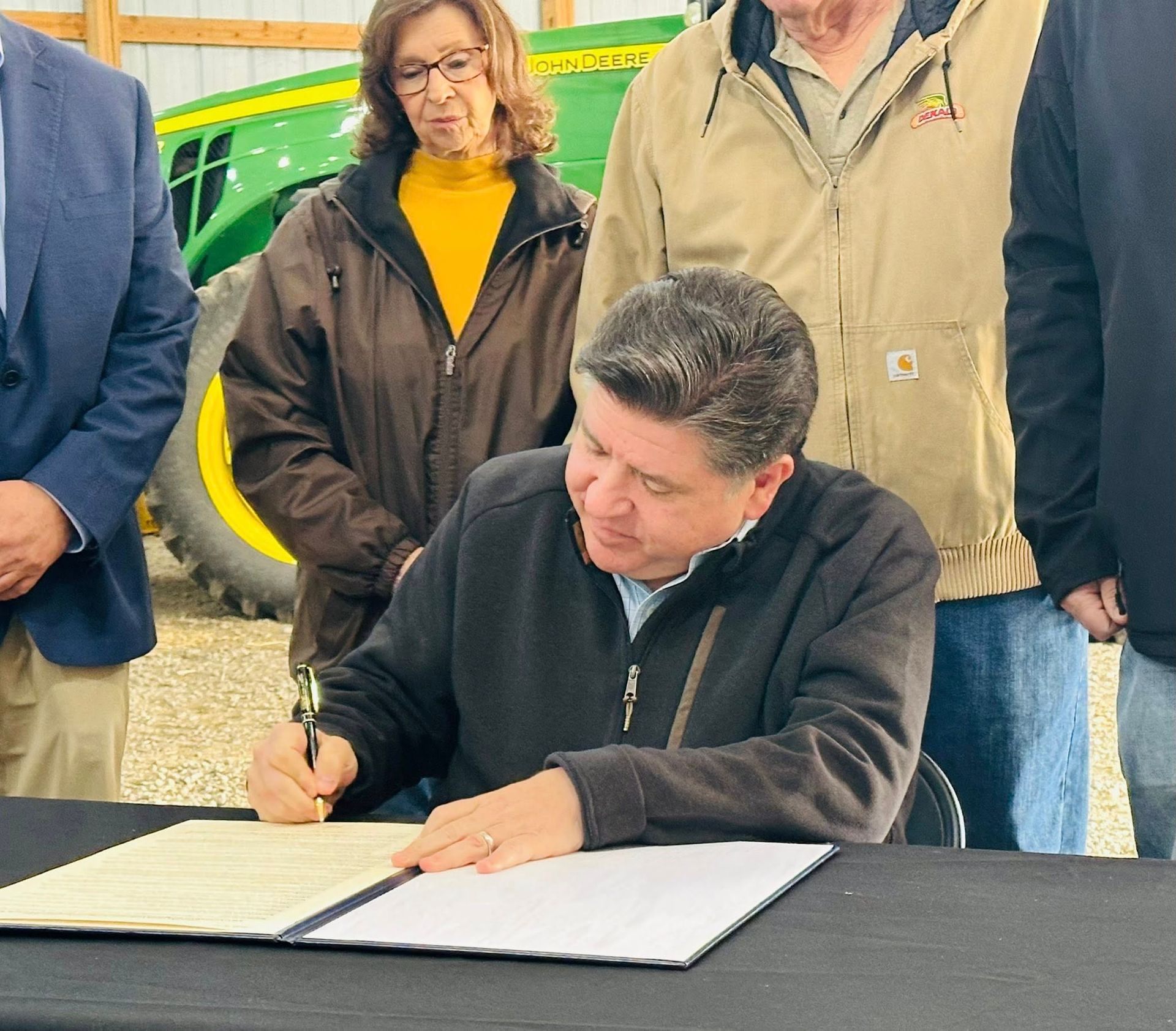 Man signing documents at a table. Others watch nearby, a tractor visible in the background.