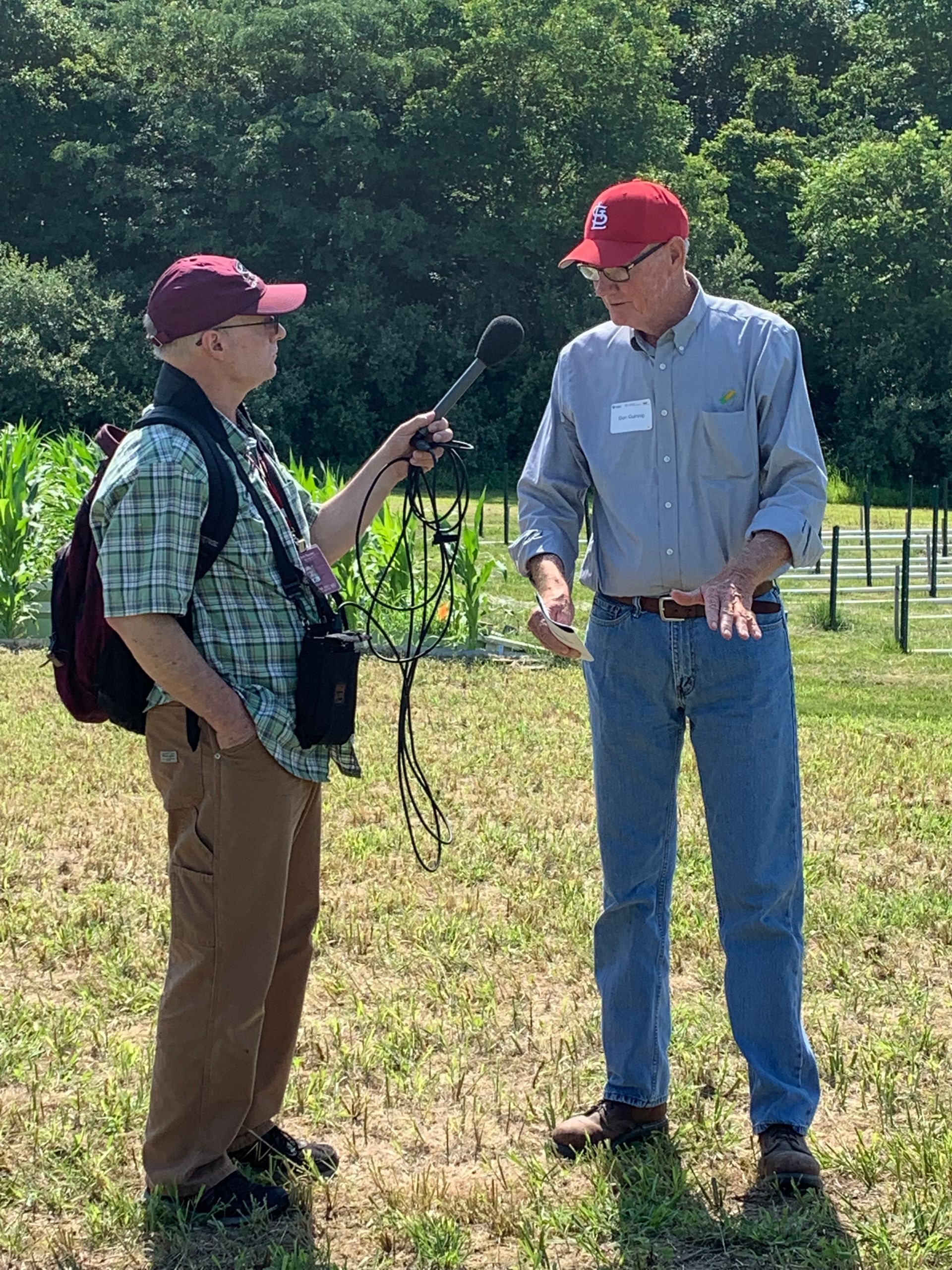 A person interviews a man in a field. The interviewer holds a microphone. Both wear hats.