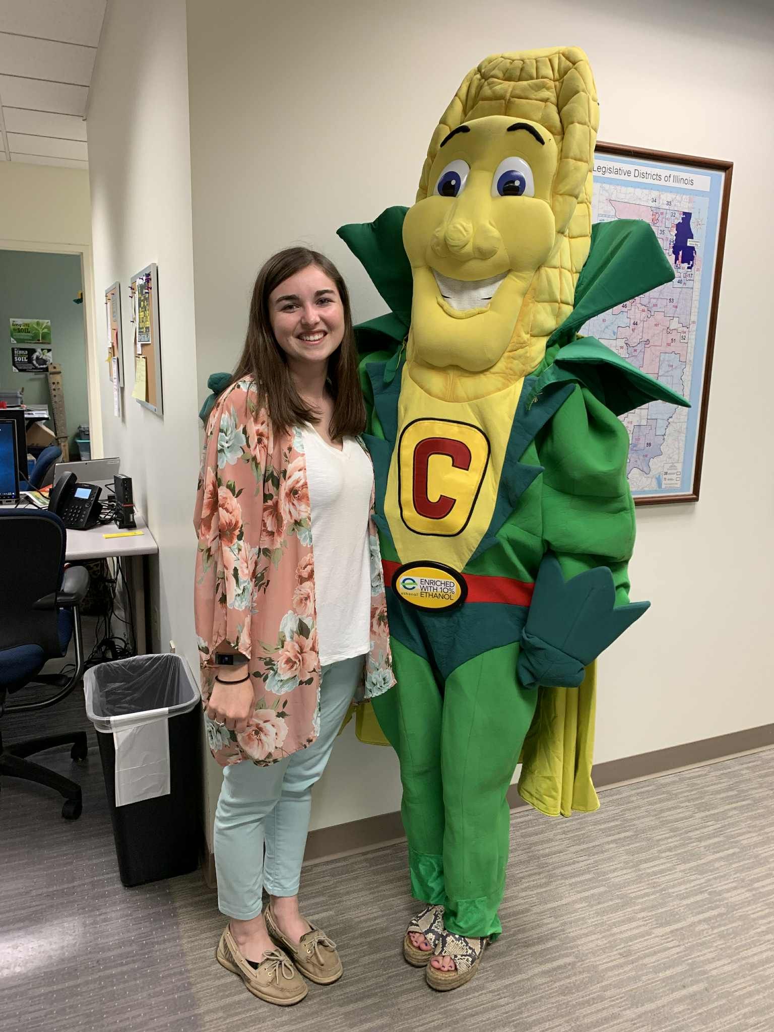 Woman in floral shirt poses with Corn superhero, in an office setting.