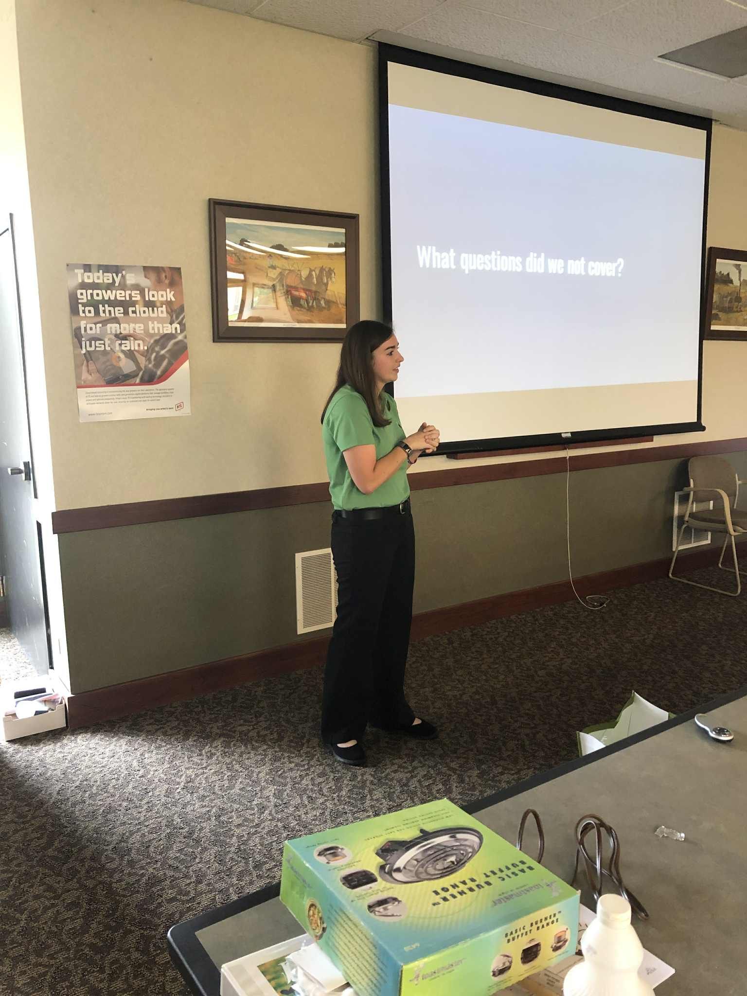 Woman giving a presentation. She stands near a screen with text, in a room with a table.