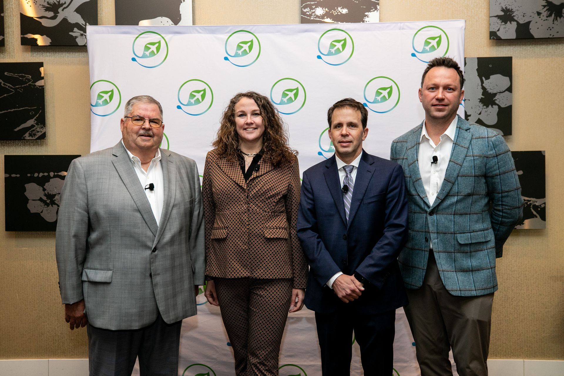 Four people in suits stand in front of a backdrop with a logo.