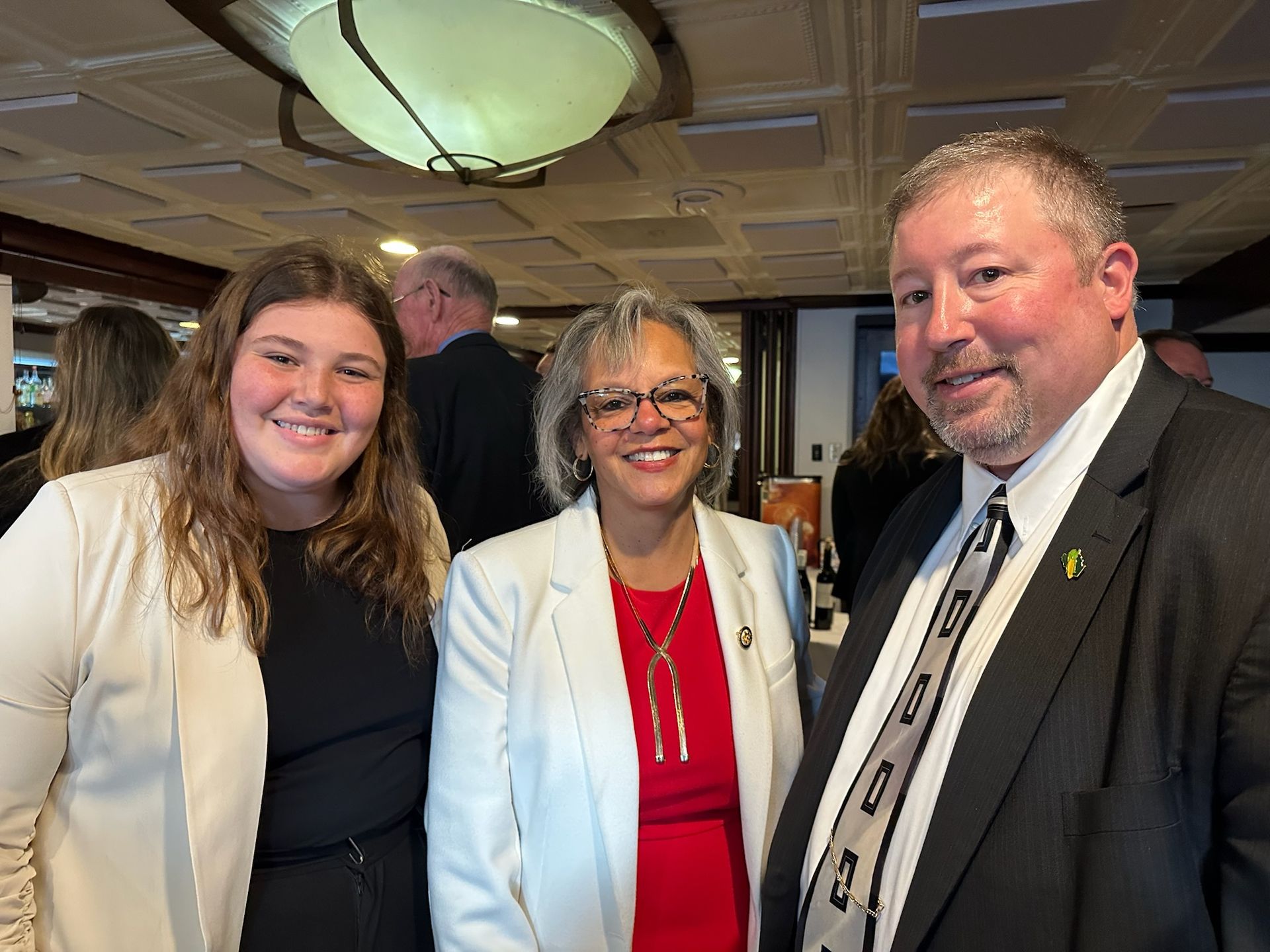 Three people smiling at a formal event, woman in red blazer and necklace, man in suit and tie.