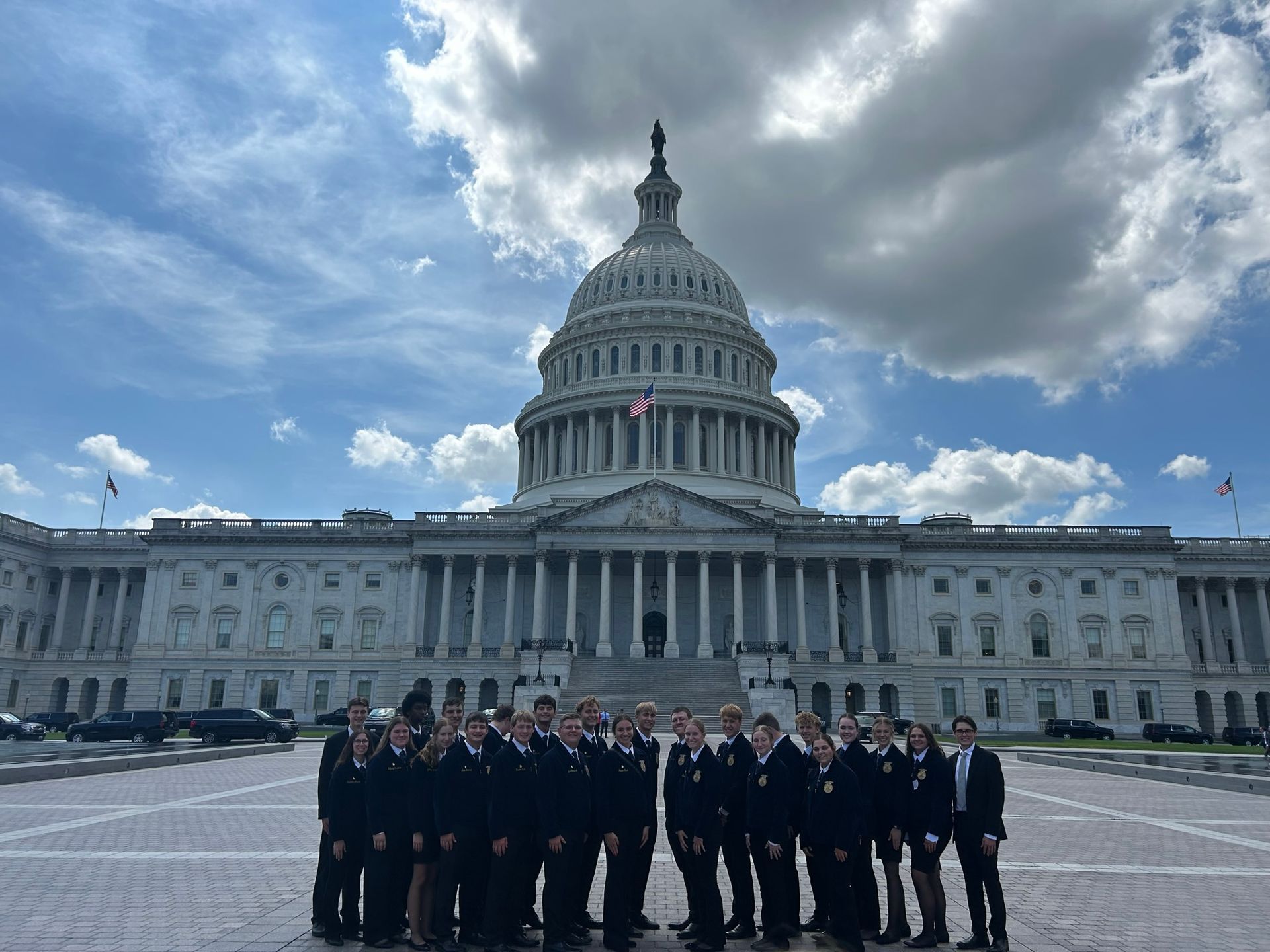 Group of people in dark suits pose in front of the US Capitol Building on a sunny day.