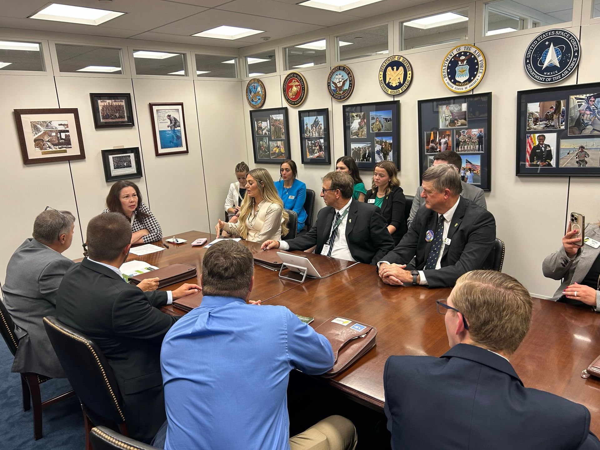 People seated around a conference table in a room with military emblems and photos on the walls.
