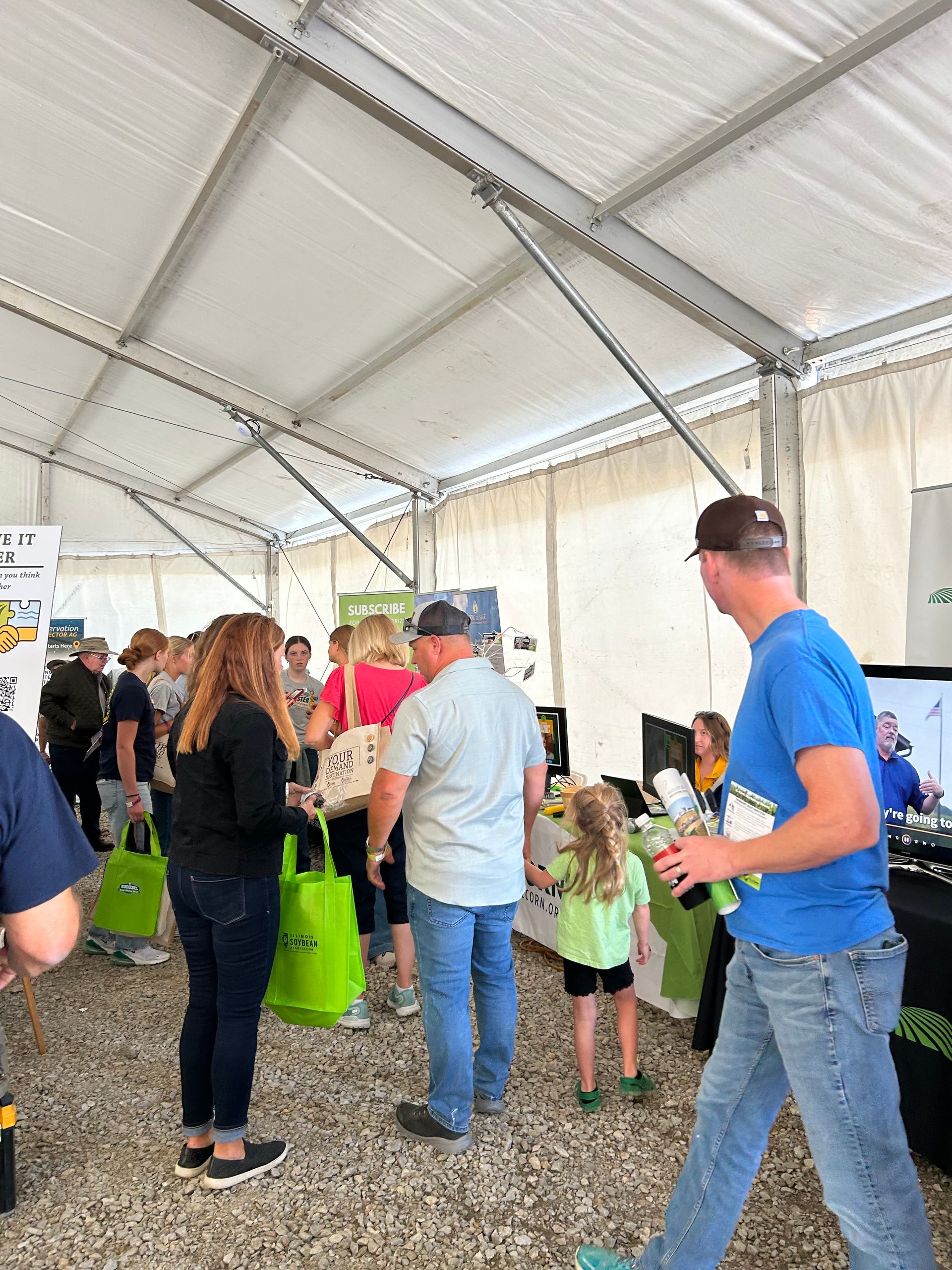 People at an outdoor event, gathered in a tent. Several adults and a child are present near a vendor display.