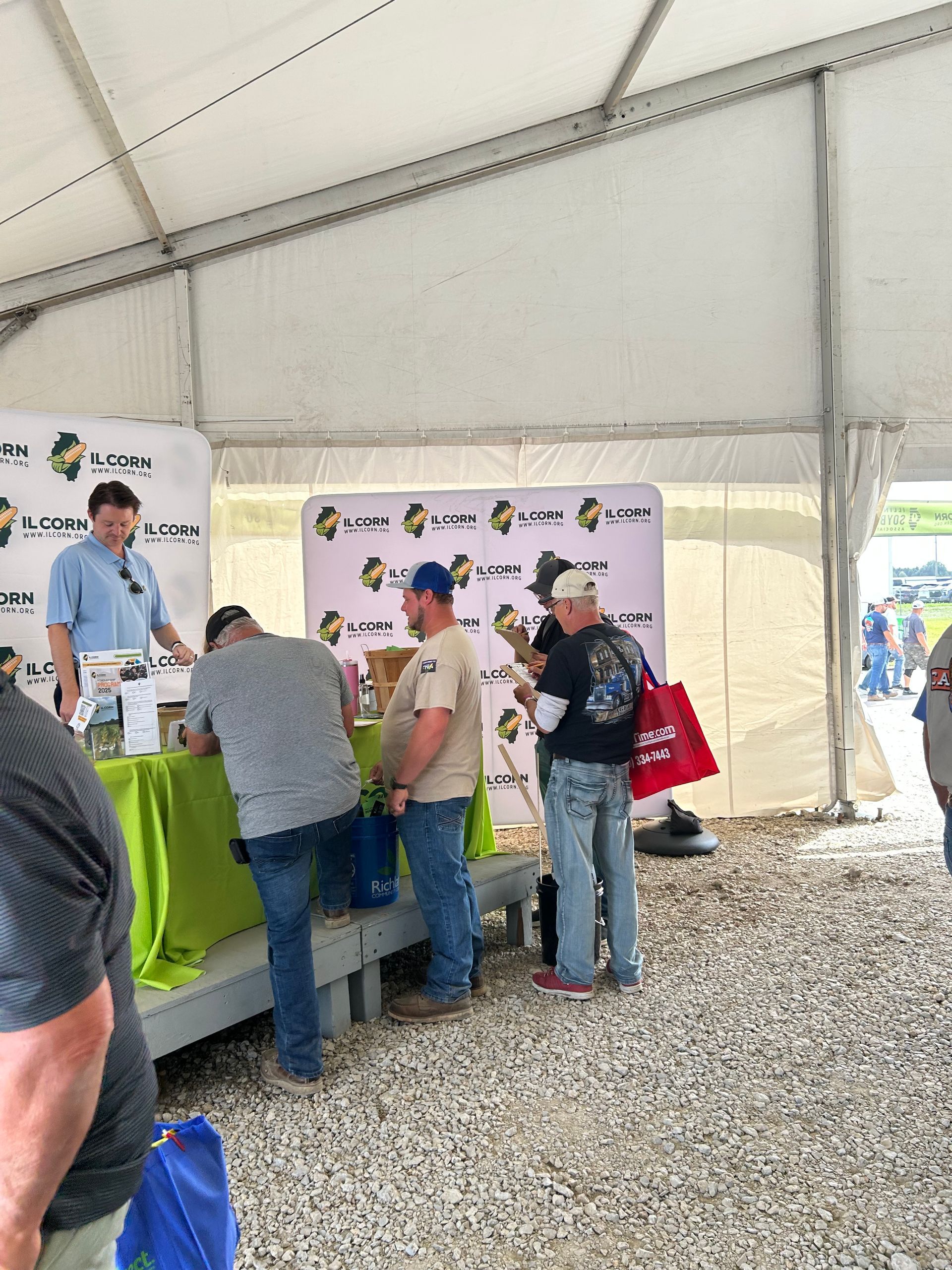 People at a tent booth, signing papers. A man stands behind a table with a logo backdrop. Outdoors, daytime.