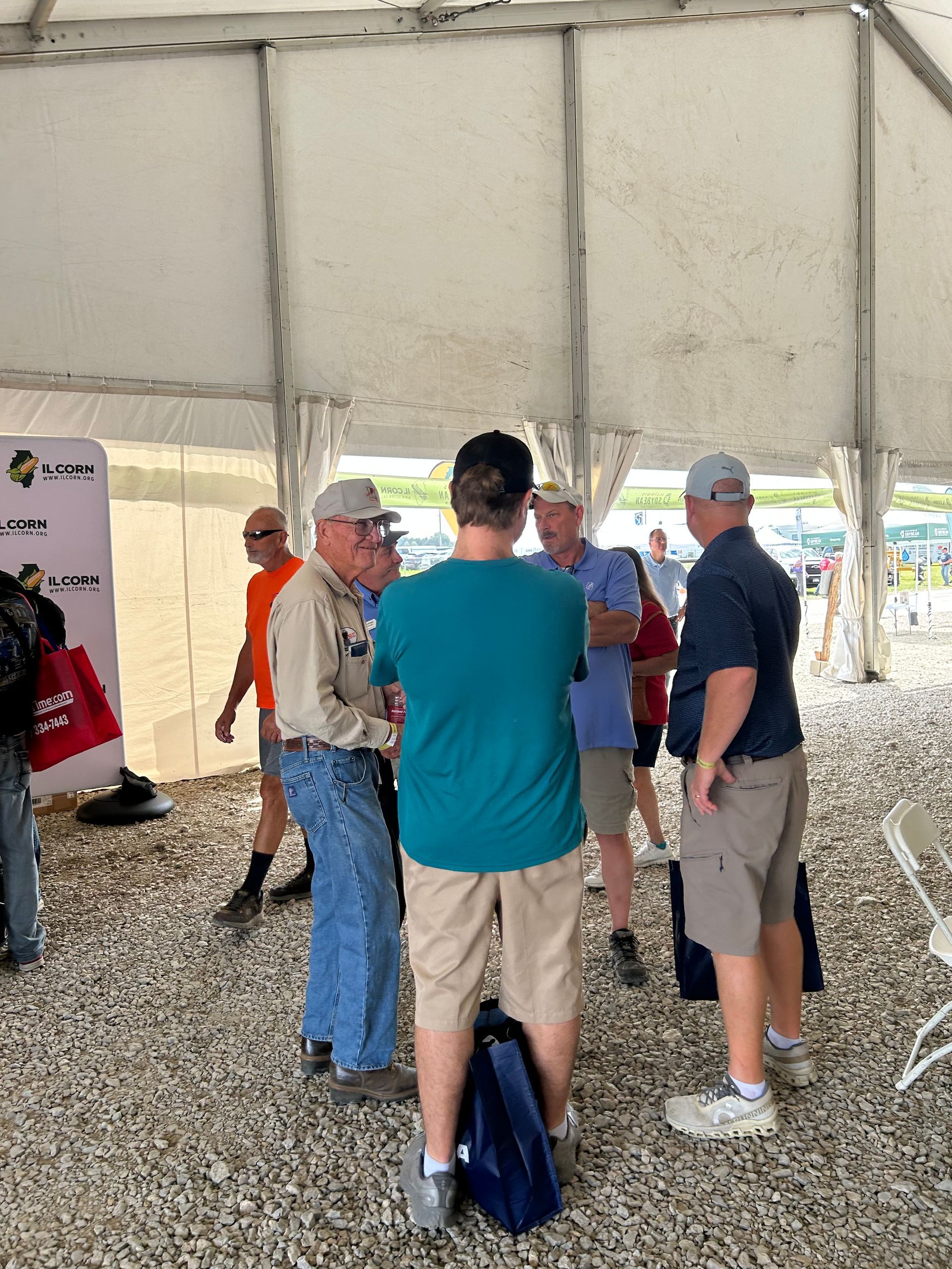People in a tent, conversing. Some are wearing hats, casual attire. Gravel ground.