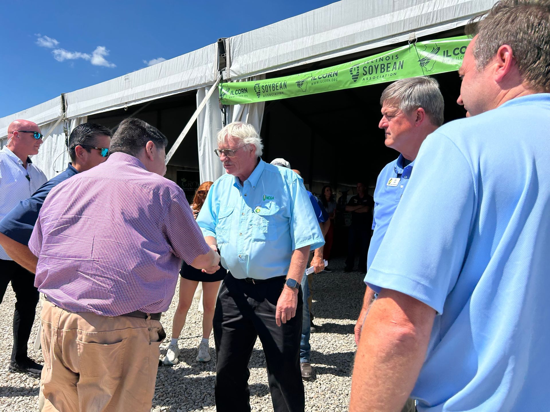 Bernie Sanders shakes hands with a man in a striped shirt outside a tent on a sunny day.