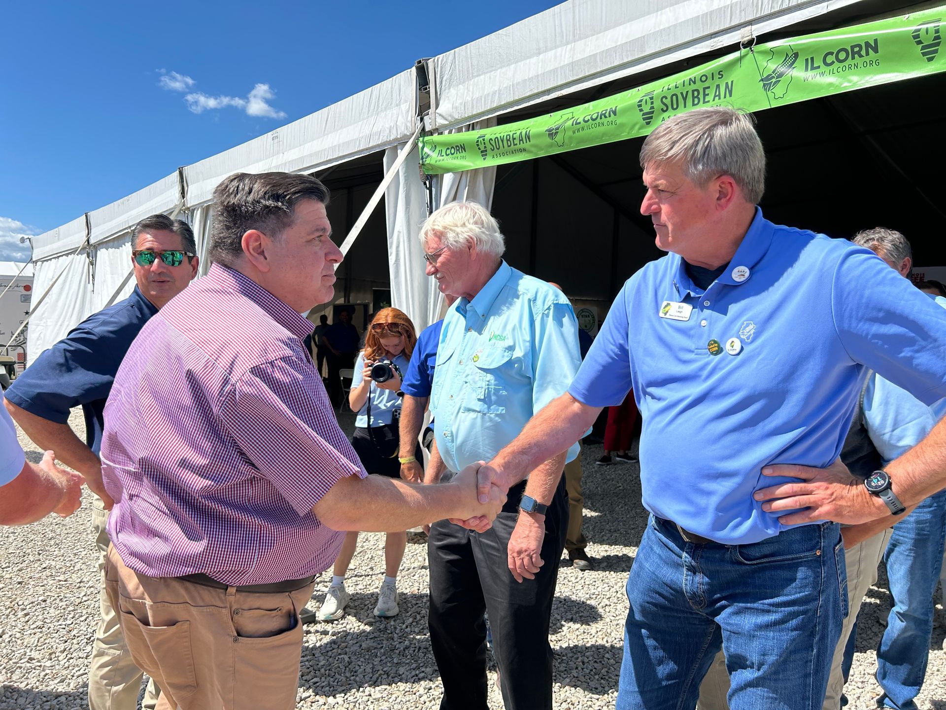 Two men shaking hands at an outdoor event. Others watch, under a tent.