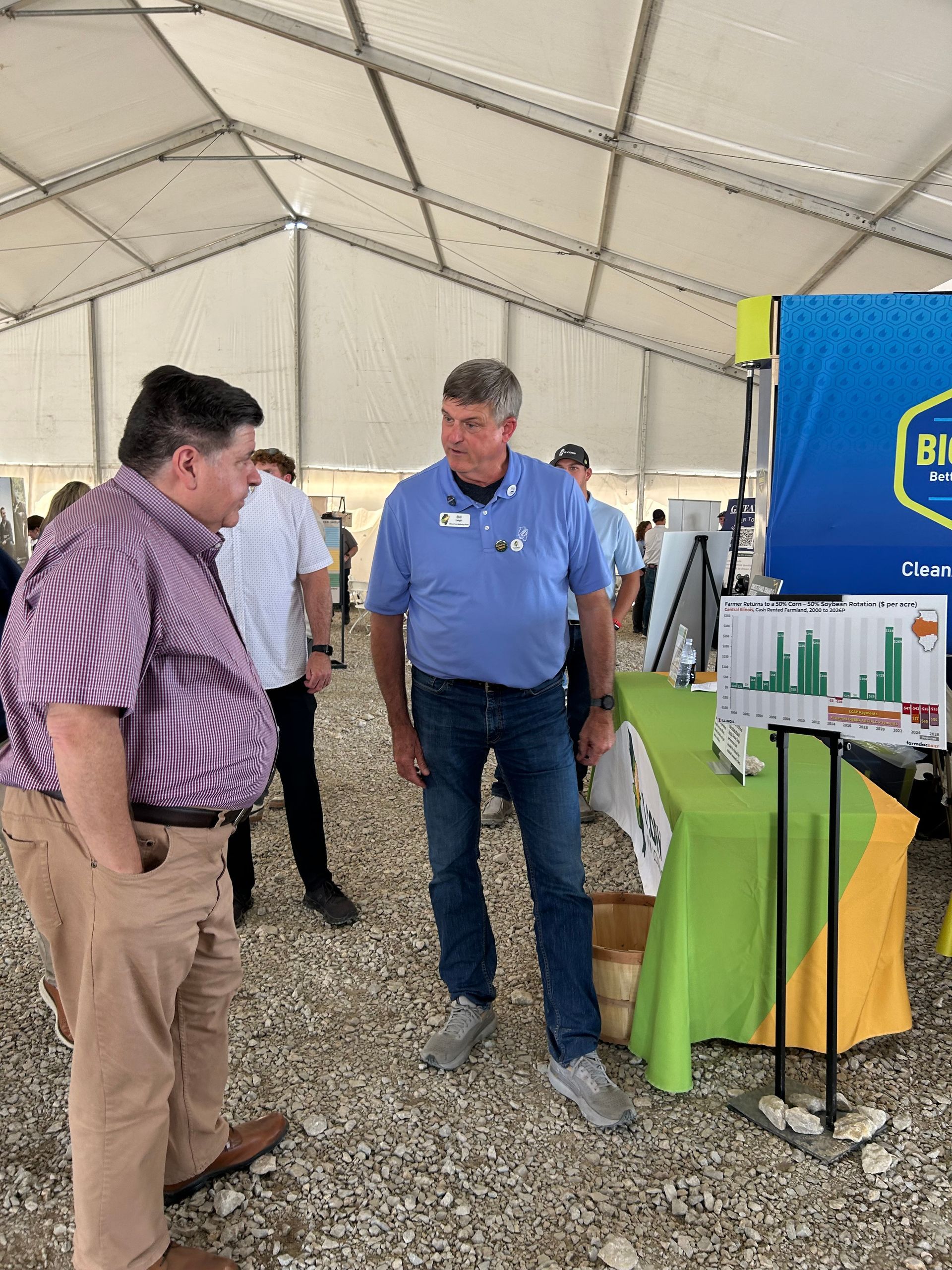 Two men talking at a trade show booth under a white tent. One wears a blue shirt, jeans. Other wears a pink shirt and khakis.