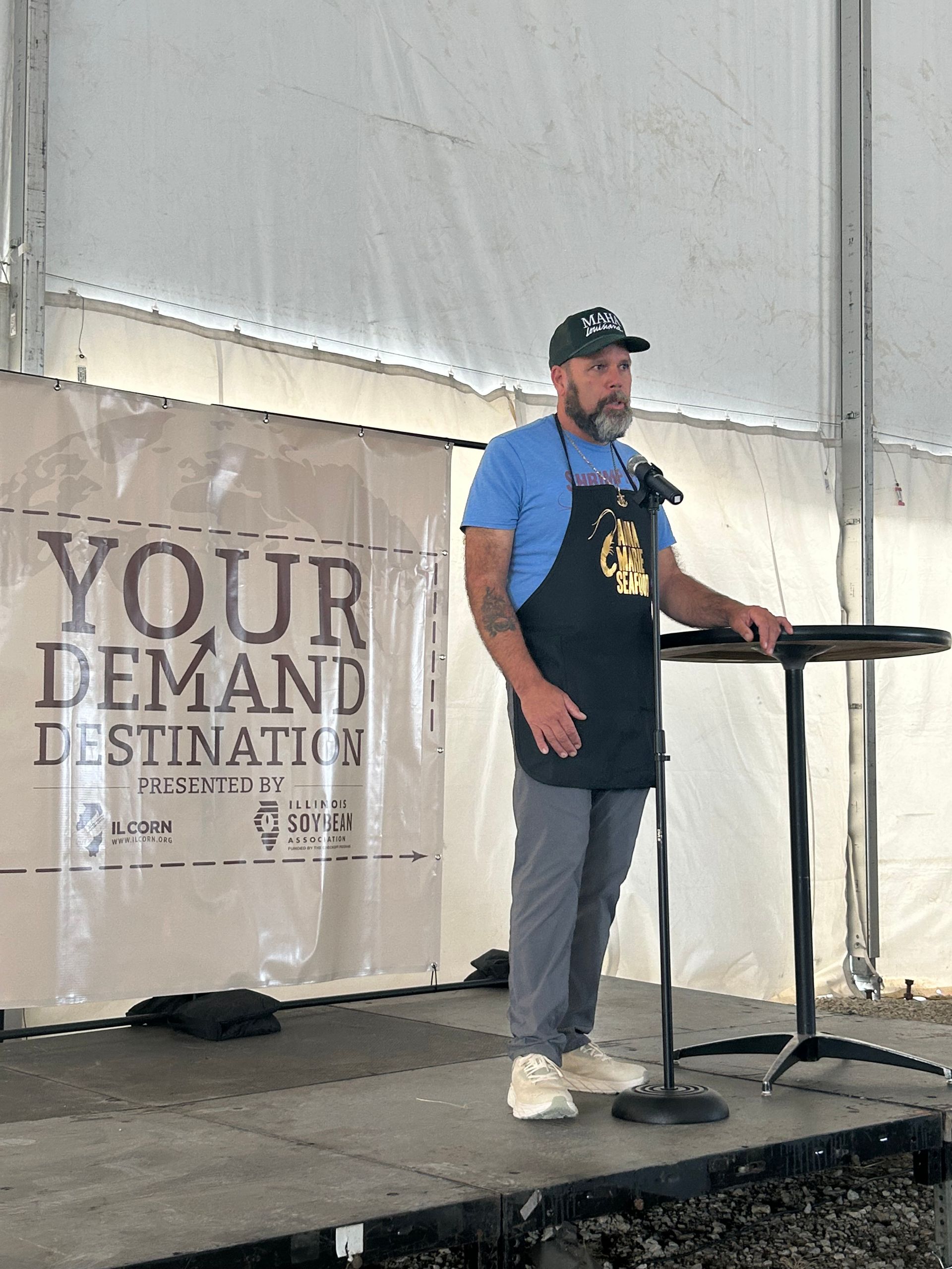 Man speaking on a stage, wearing a black apron and baseball cap,