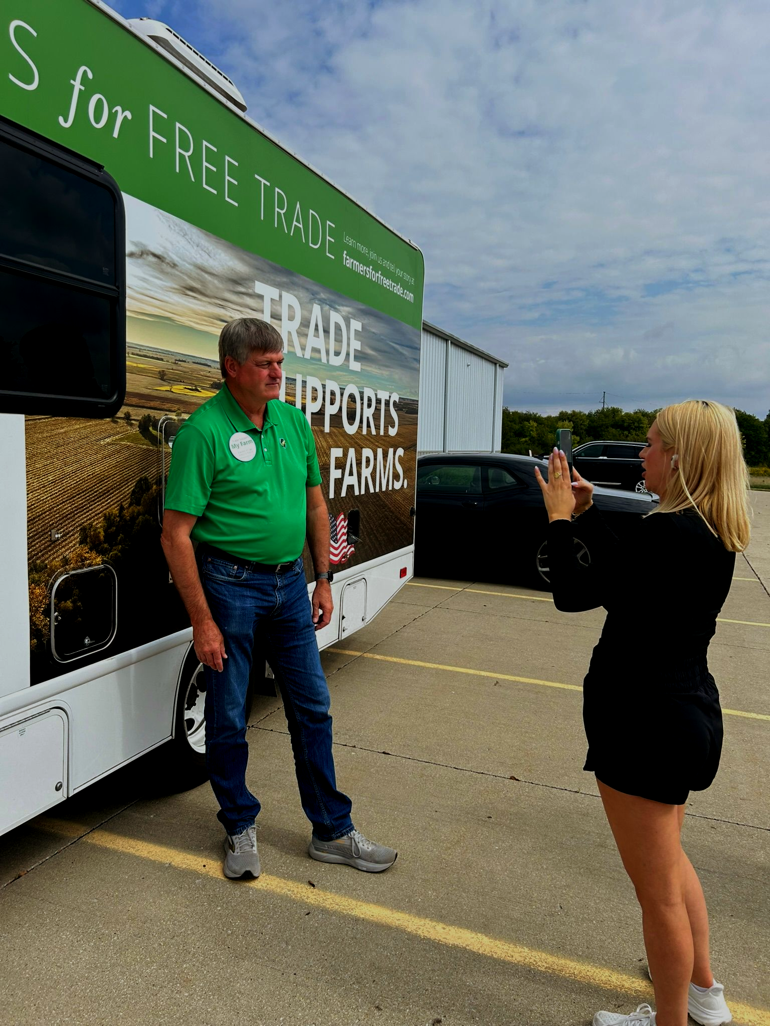 Man poses for photo next to bus promoting free trade; woman takes photo with a phone outside.