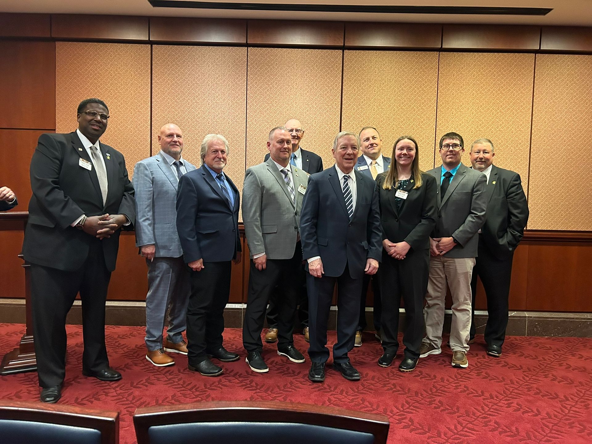 A group of people in business attire stand in a row for a photo in a wood-paneled conference room with red carpet.
