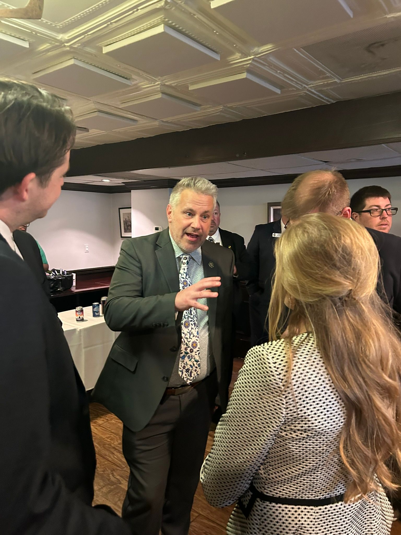 A group of people in business attire networking in a room with a paneled ceiling, as one person gestures while speaking.