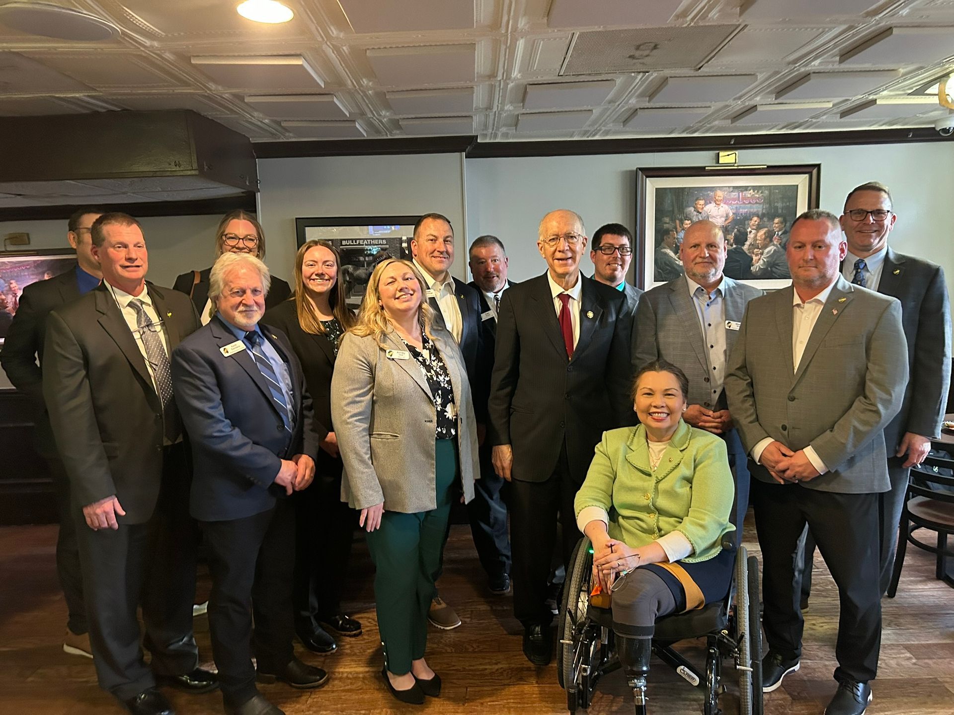 A group of professionals in business attire stand and pose for a photo indoors, with one person seated in a wheelchair.