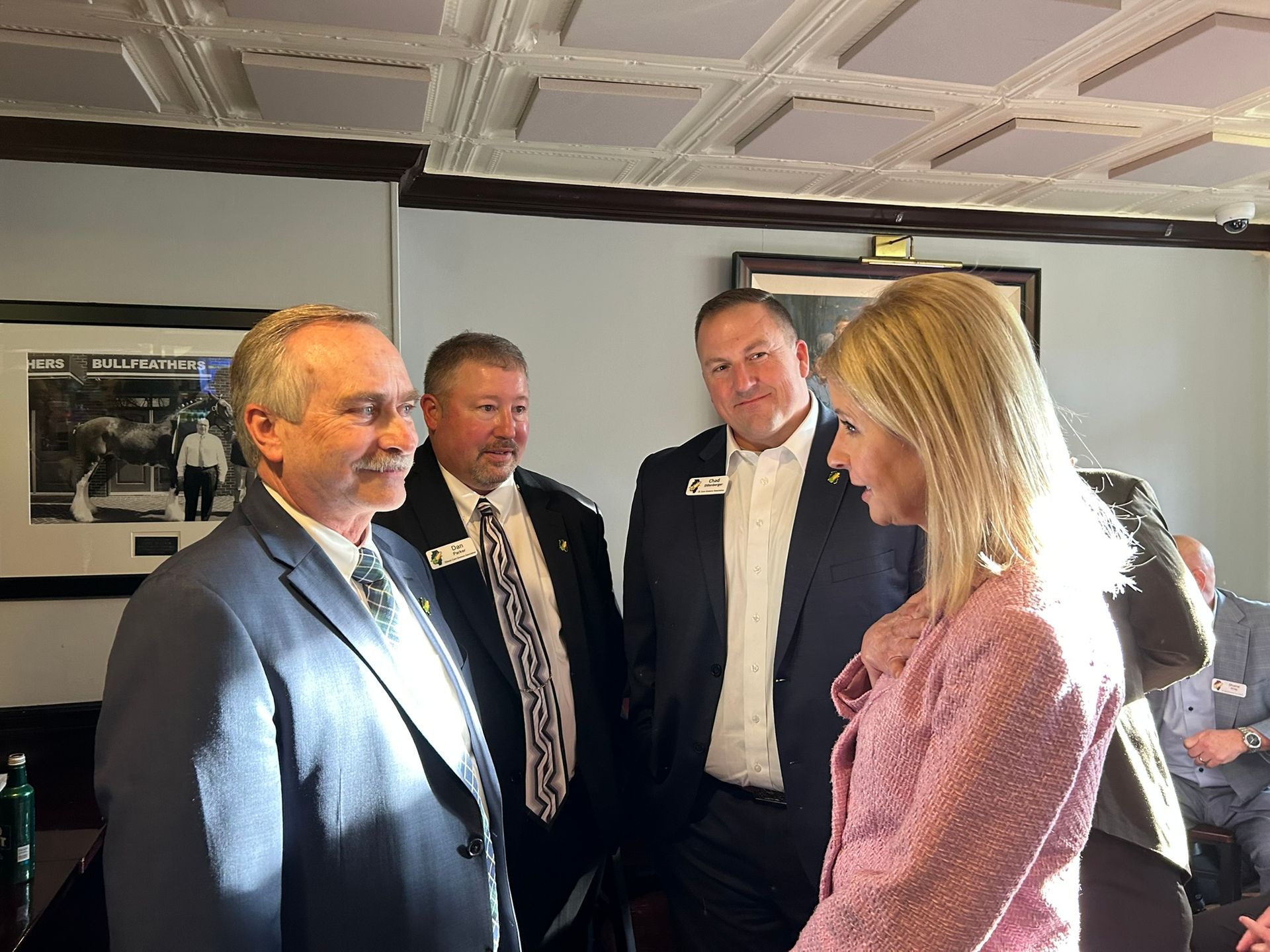 Four people talk in an indoor space with a decorative ceiling; one wears a pink jacket and faces the others.