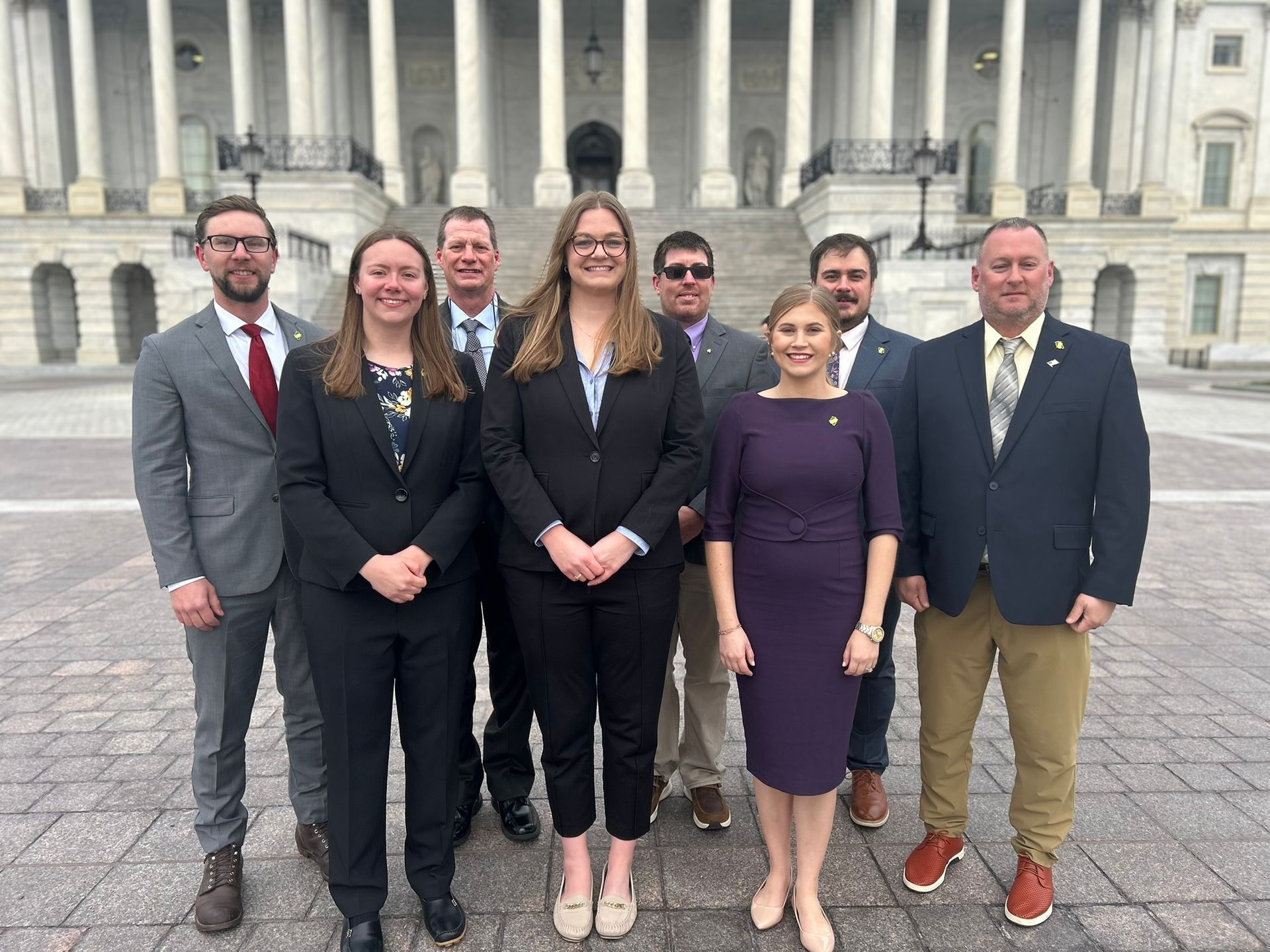 A group of eight people stand in professional attire in front of the US Capitol building.