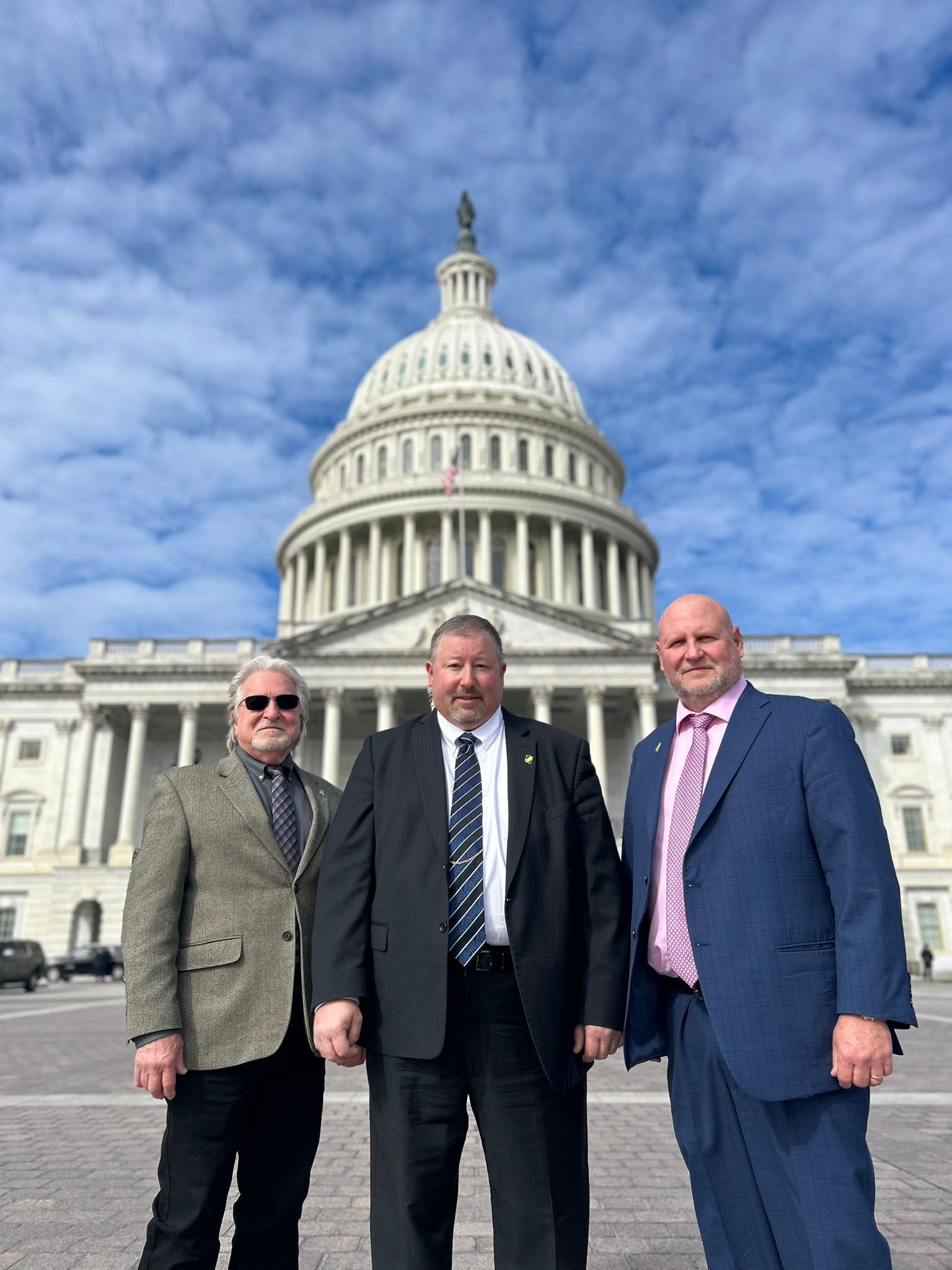 Three men in suits stand in front of the United States Capitol building under a partly cloudy blue sky.