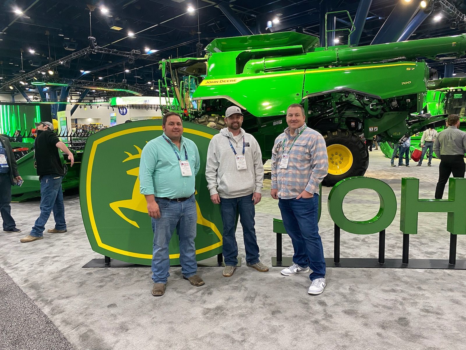 Three men pose in front of a John Deere logo and combine harvester at a trade show.