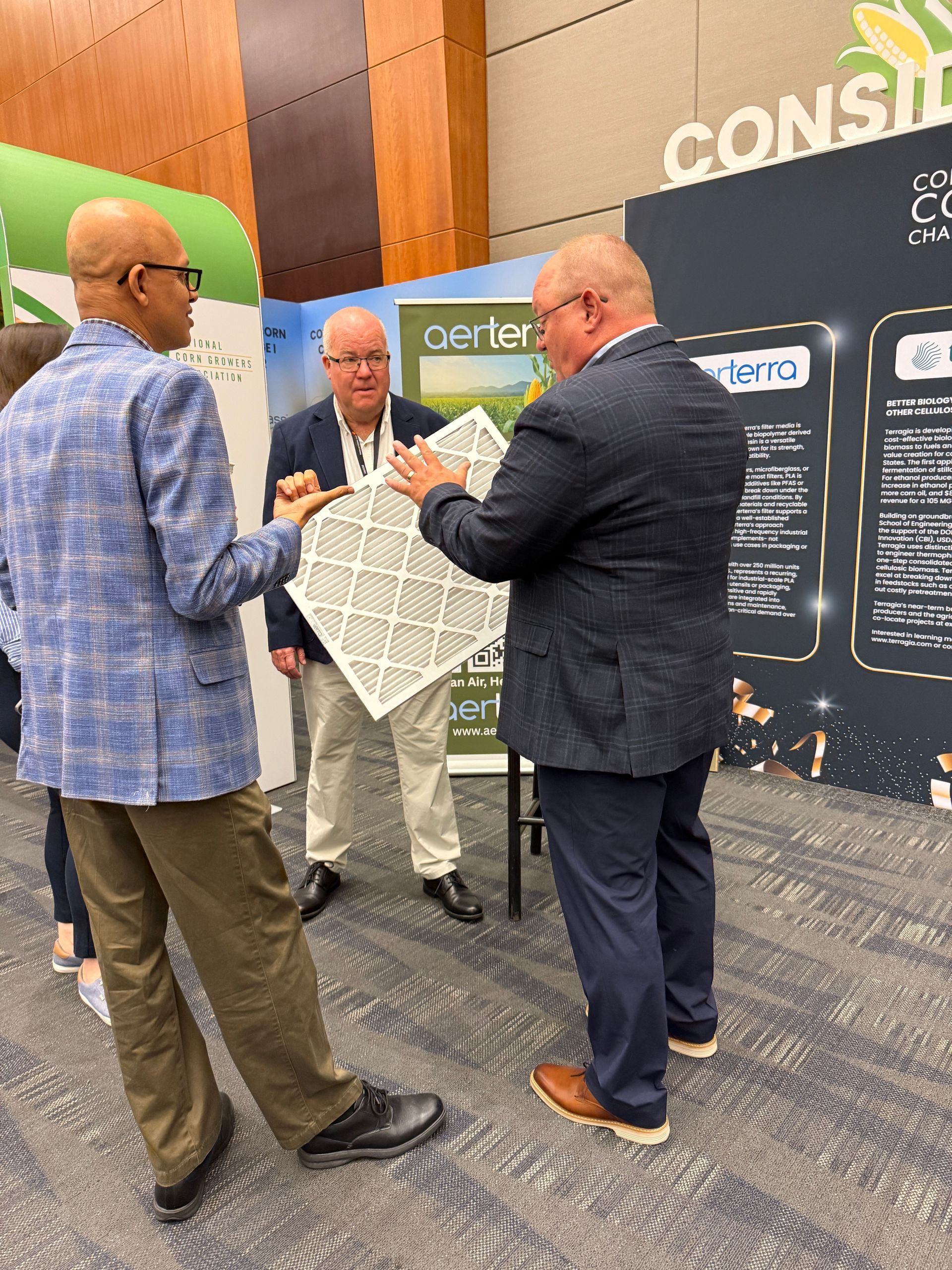 Three men examine a patterned tile at a convention booth. One points, one holds it, one looks on.