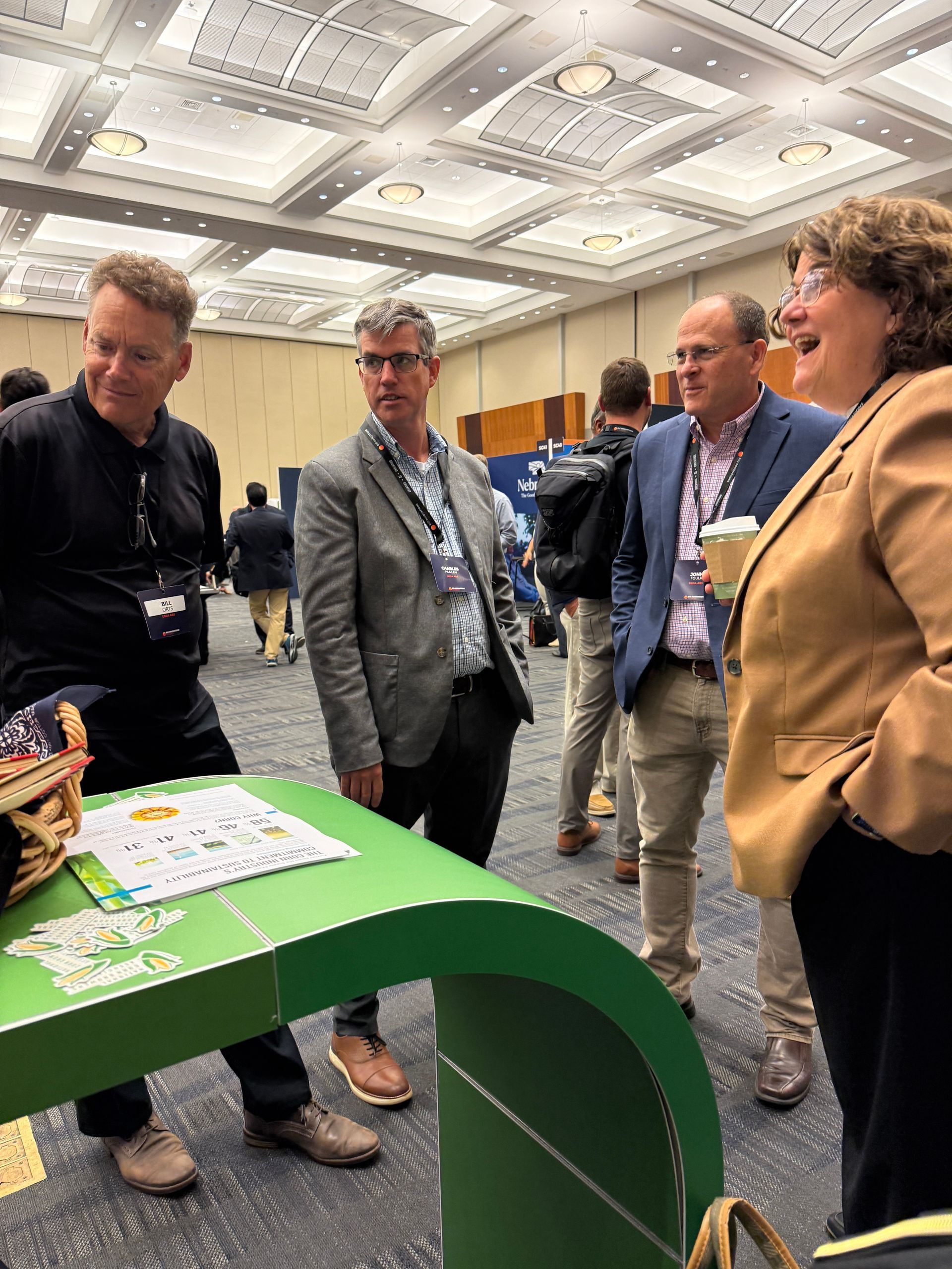 Four people conversing at a conference. Men and a woman standing near a green display.