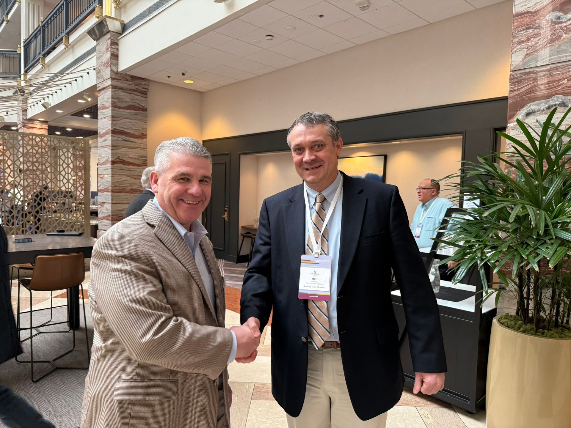 Two men shaking hands in a hotel lobby. One wears a patterned blazer, the other a navy blazer. Both smile.
