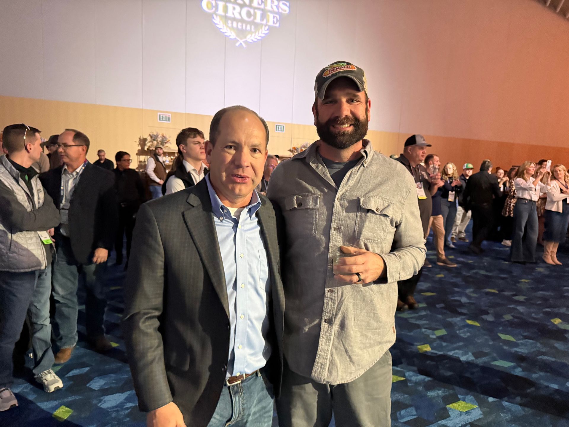 Two men posing, smiling, inside a crowded event space, banner reads 