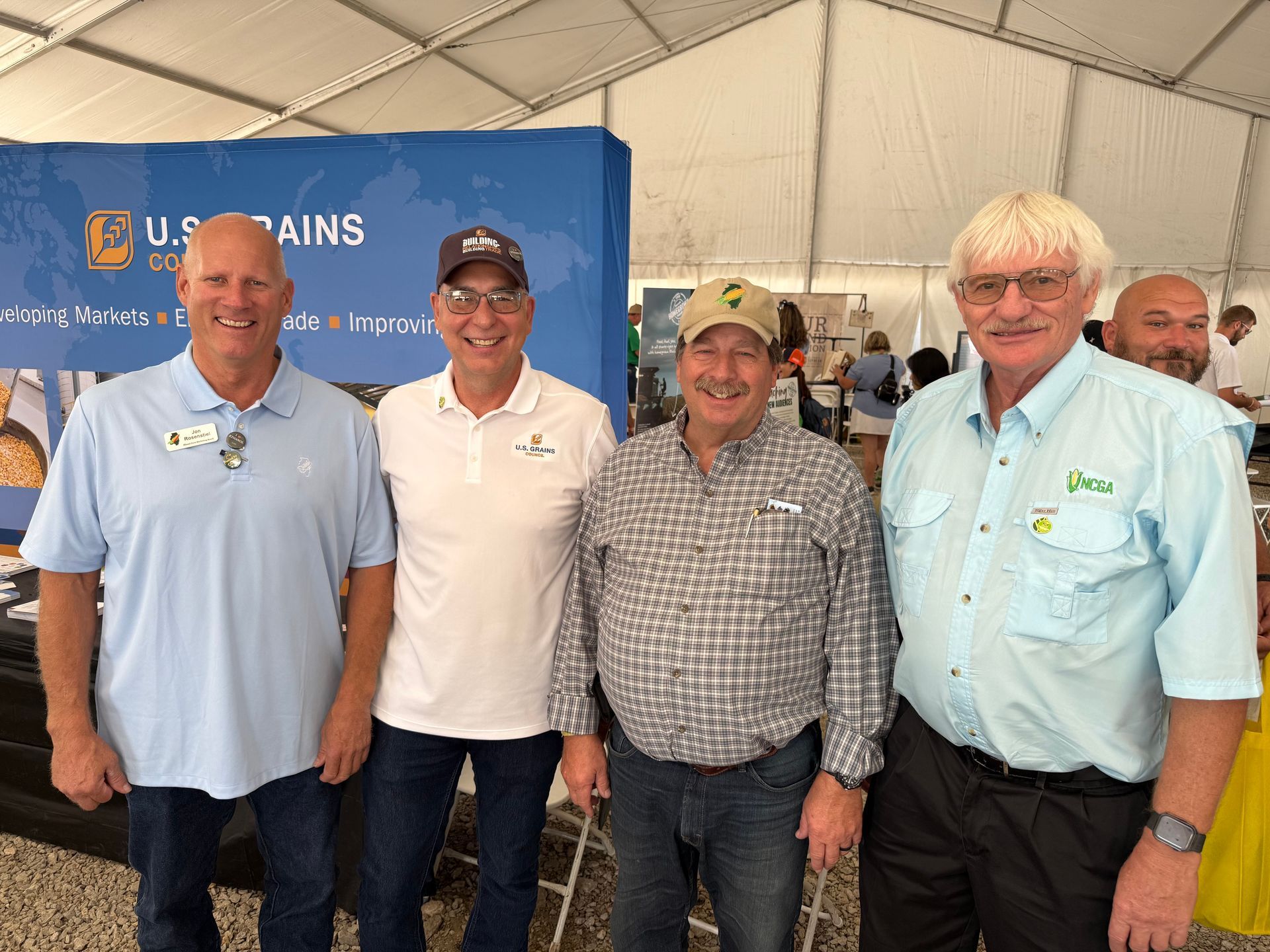 Four men smiling, under a tent. Two are wearing blue shirts. The background shows a booth with a logo.