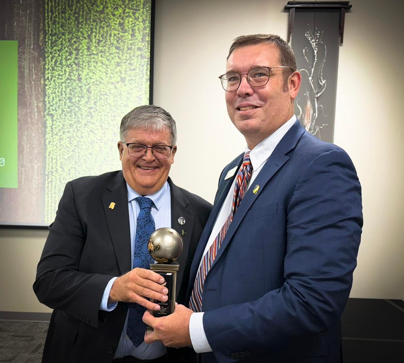 Two men, one in a suit, the other in a suit jacket, holding an award in a room with a presentation screen.