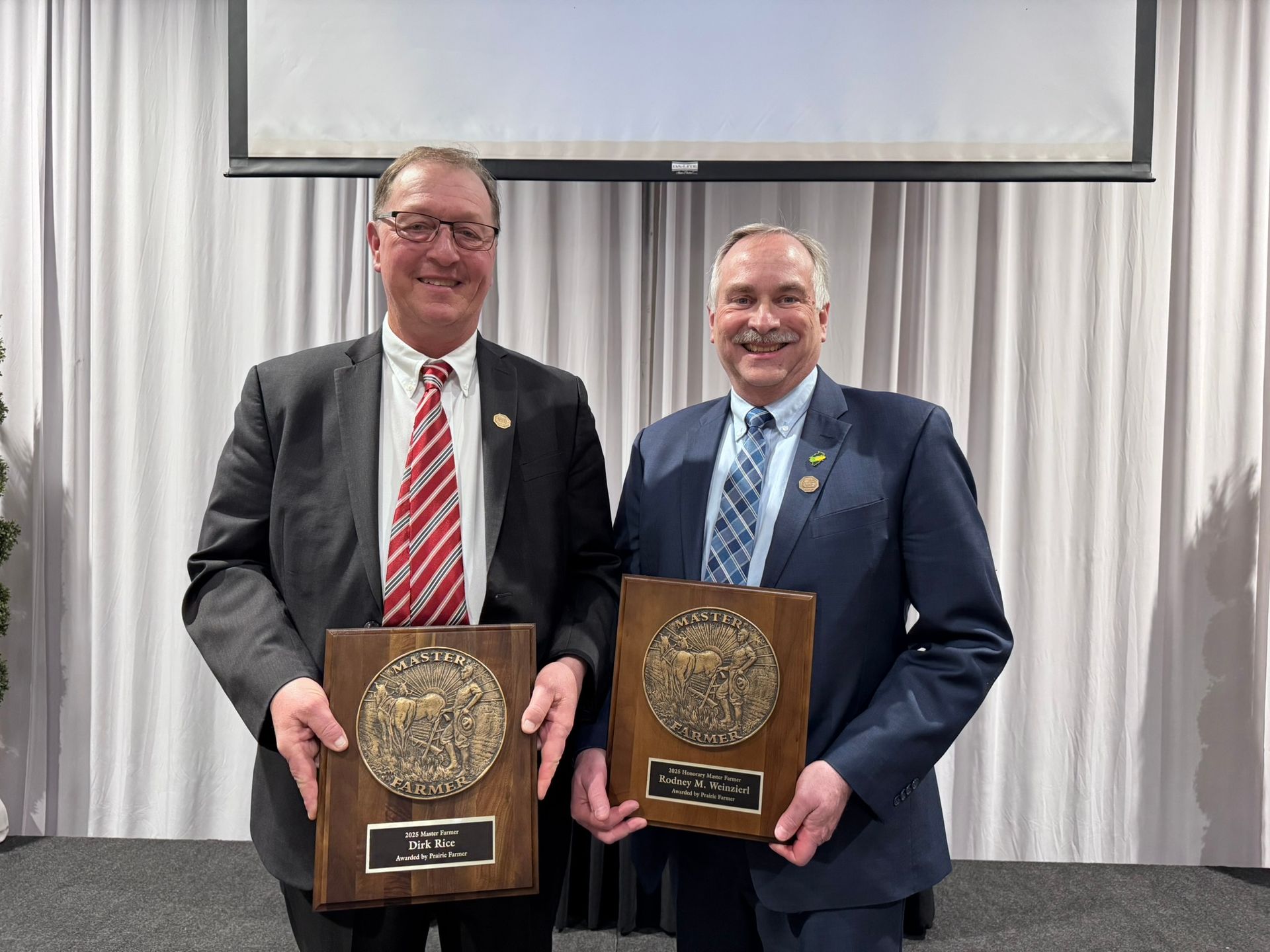 Two men holding plaques, smiling in front of a curtain. One wears a red tie, the other a blue tie.