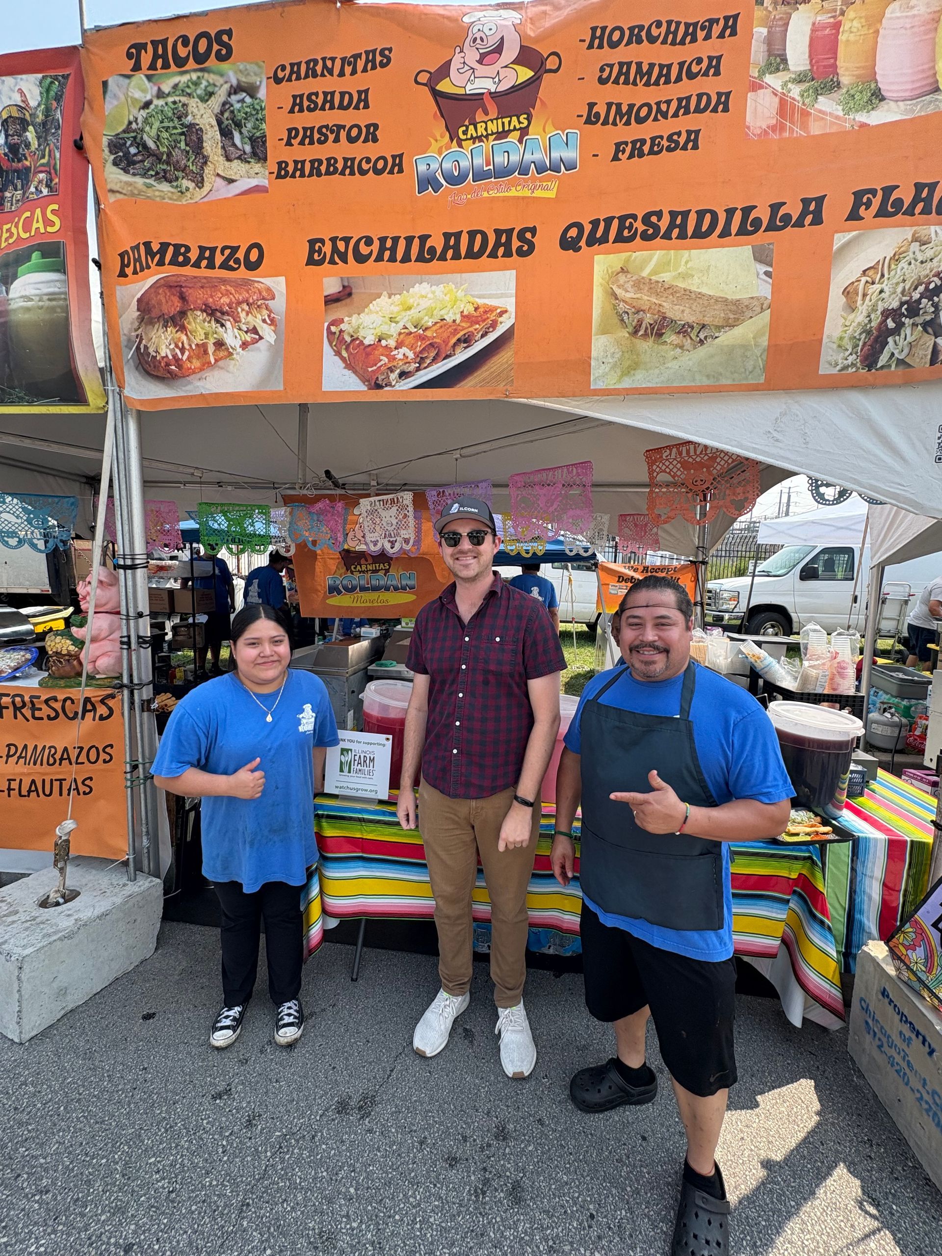 Three people at a food stand with a banner for Mexican food. They are smiling.