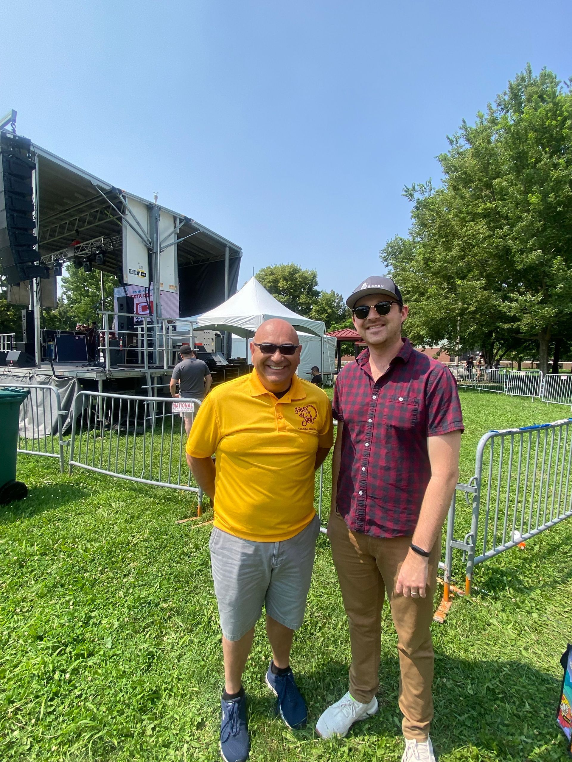 Two men standing on grass in front of a stage. One wears yellow shirt, shorts; the other, a button-down shirt, pants, and a hat.