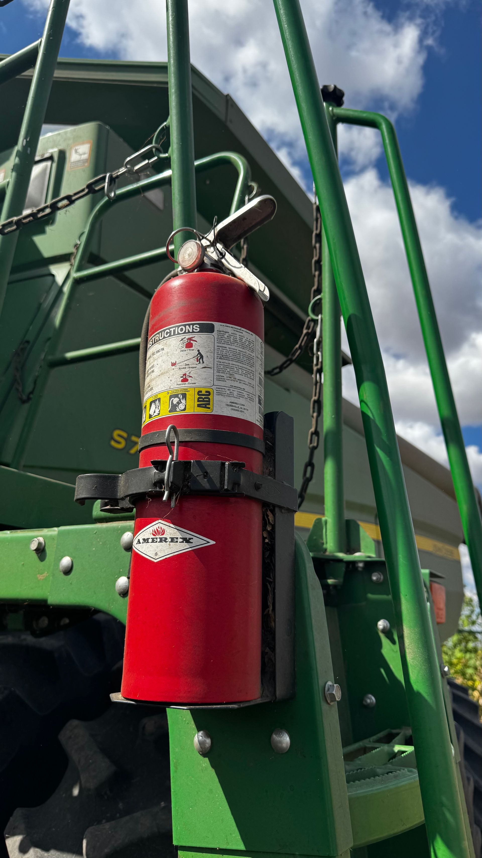 Red fire extinguisher mounted on green farm equipment, near a ladder, under a blue sky.