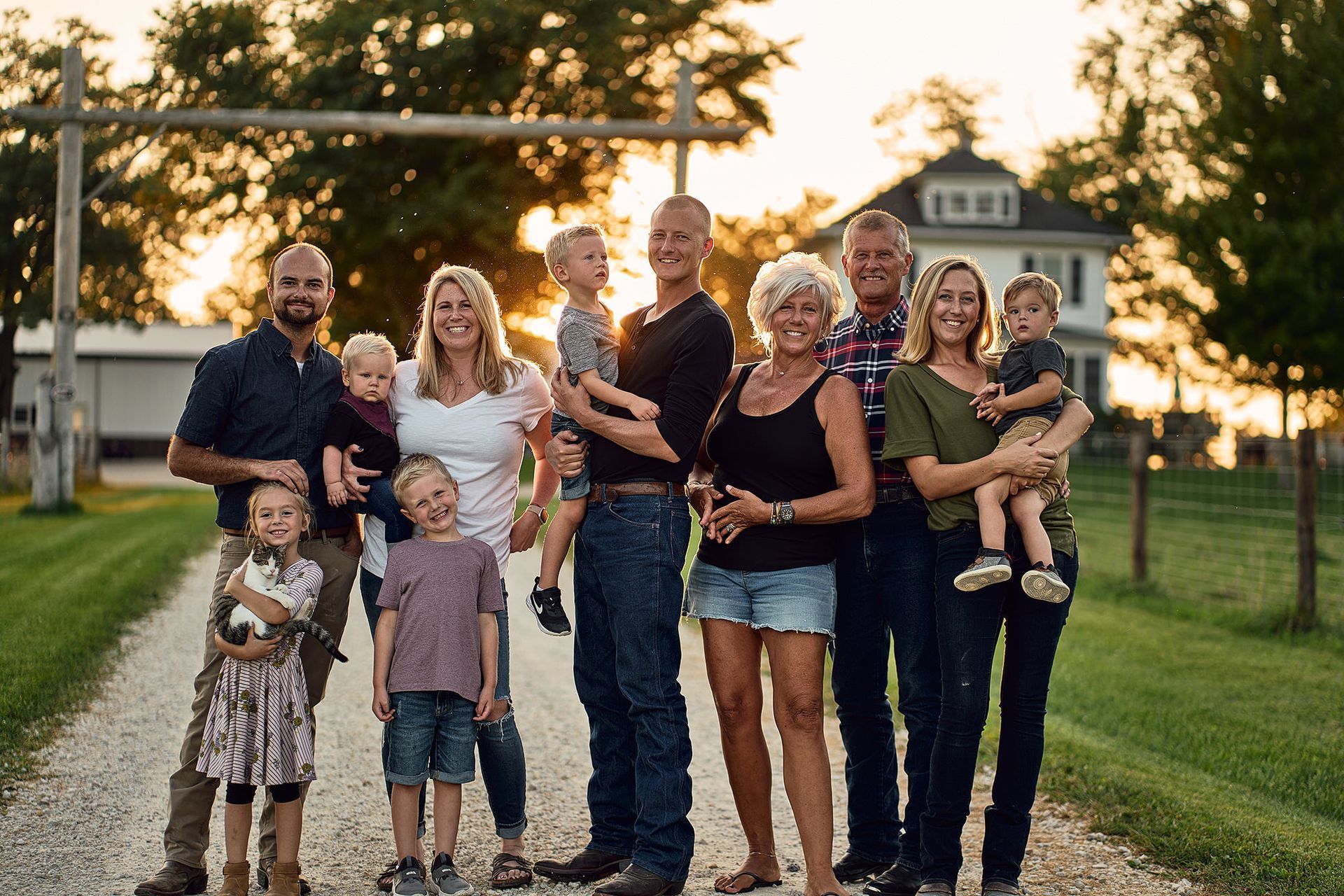 Family portrait on a gravel driveway at sunset. People smiling, holding children, and standing in front of a farmhouse.