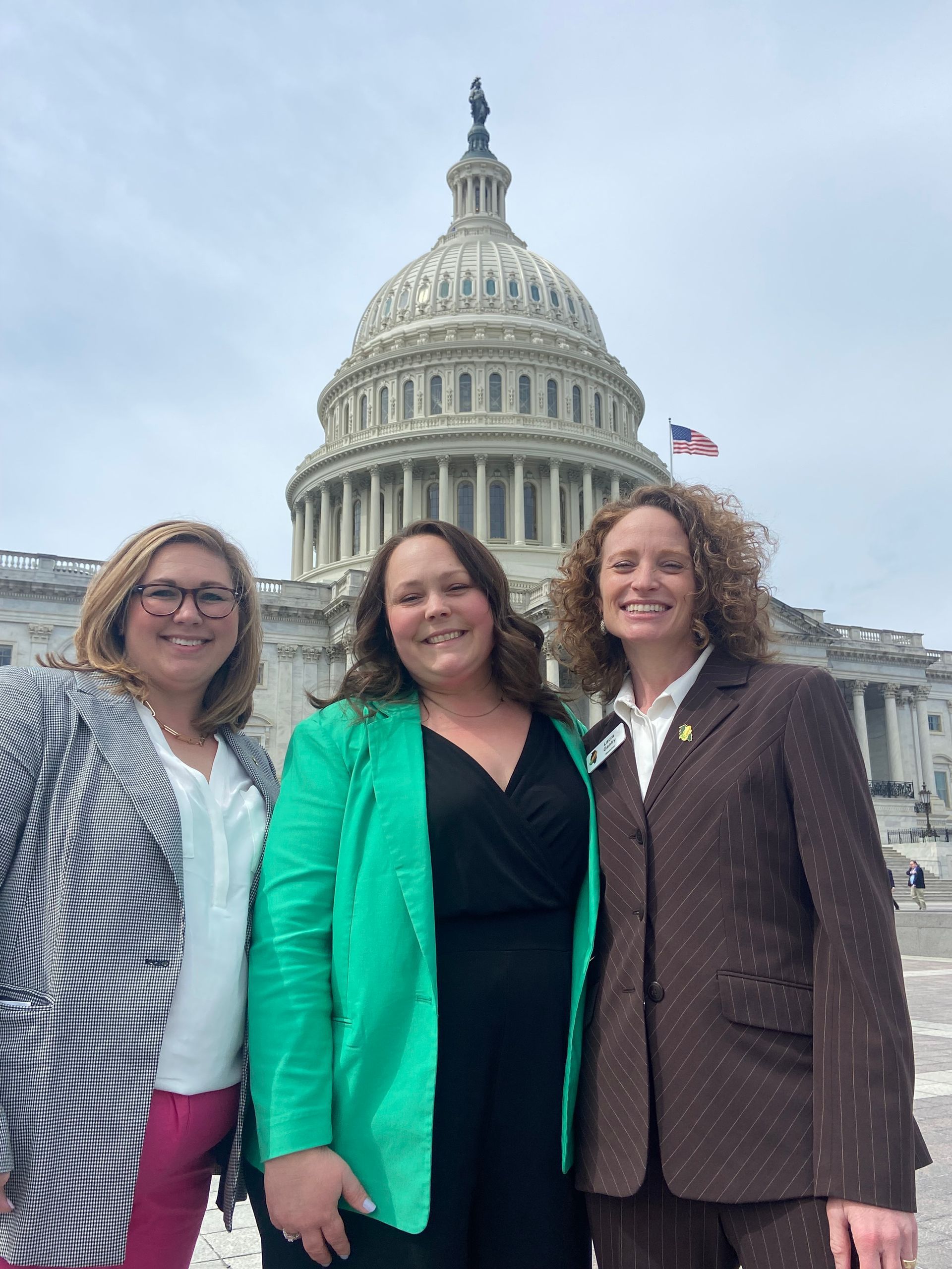Three women pose in front of the U.S. Capitol building; one wears a green jacket, others blazers.