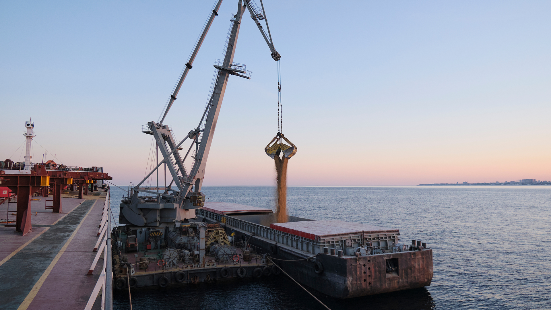 Crane loading grain onto a ship at a dock, setting sun over the water.