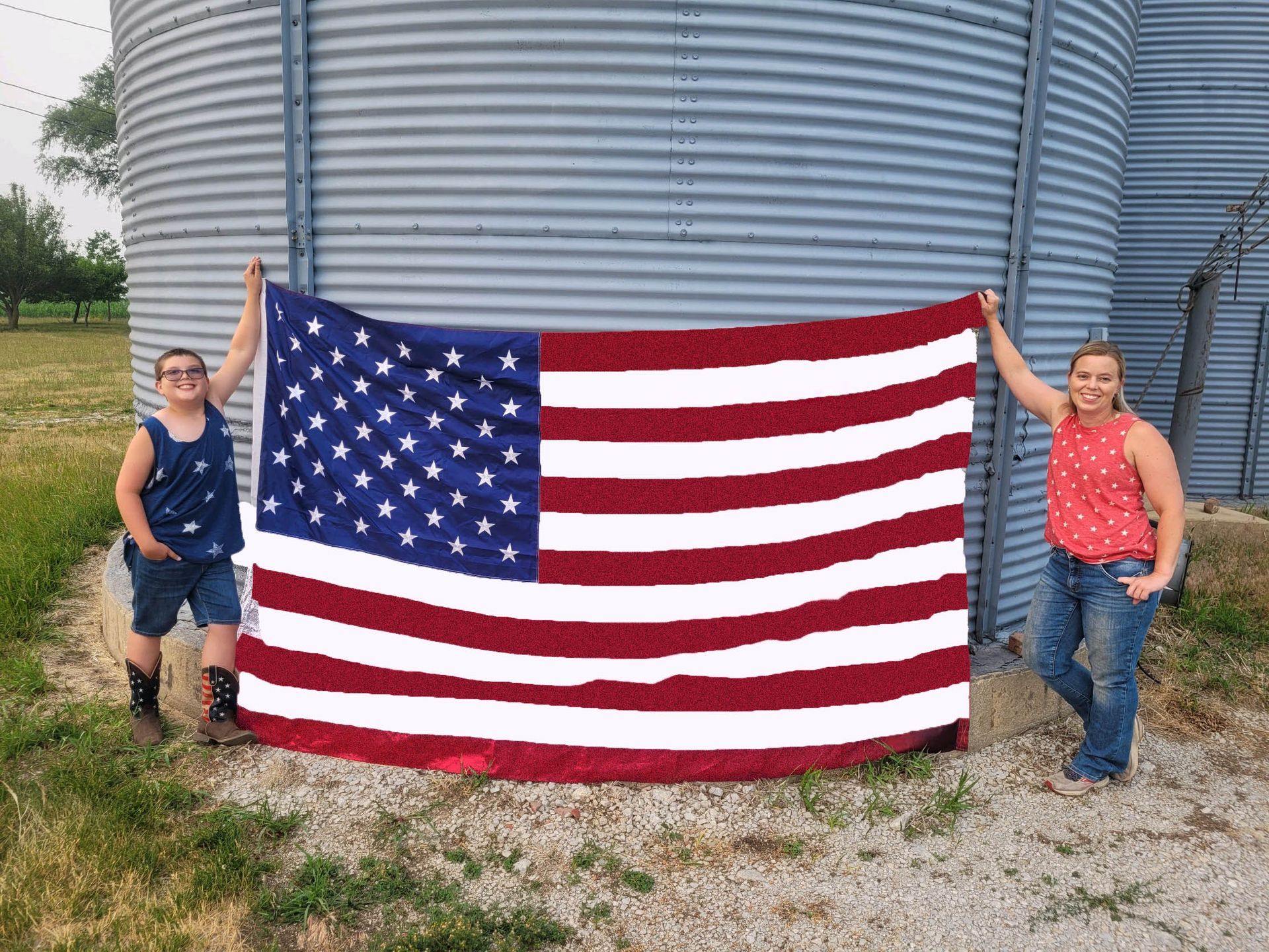 A boy and woman hold up a large American flag in front of metal silos.