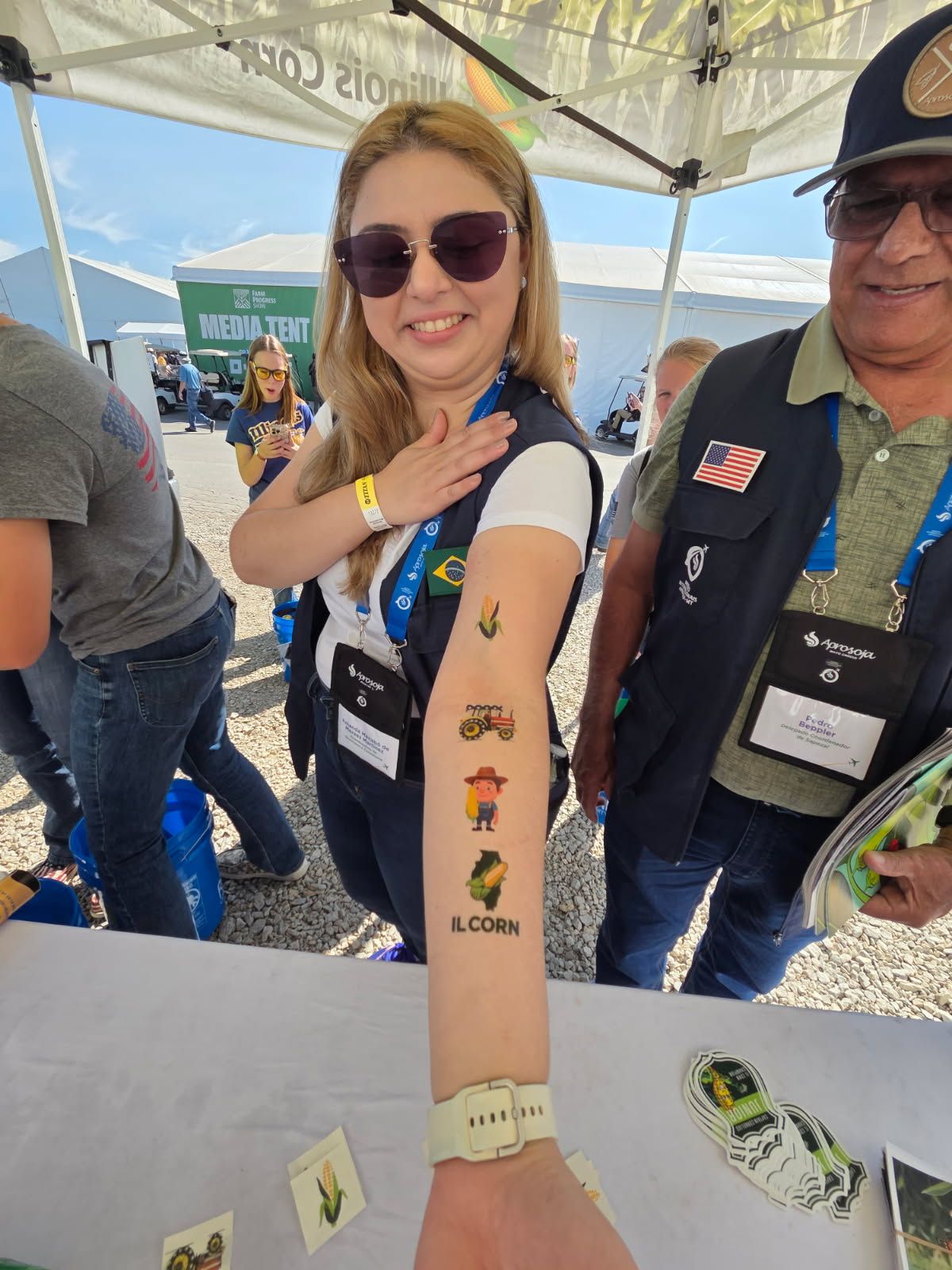 Woman with temporary tattoos smiles, standing with a man at an outdoor event; sunny day.